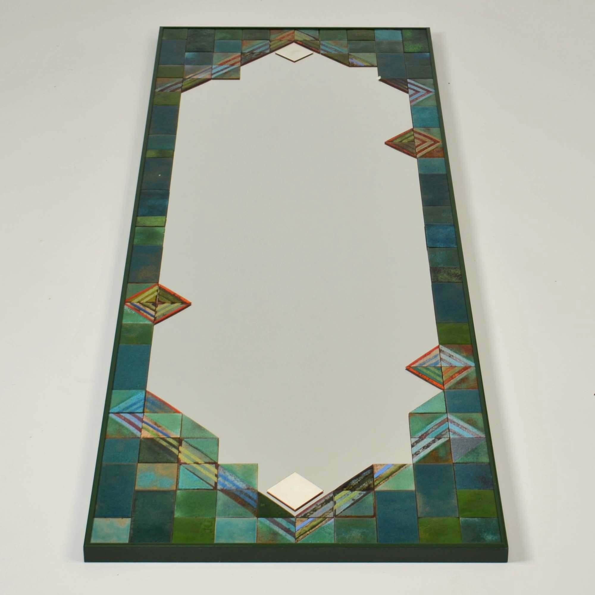 Ce miroir mural rectangulaire unique en son genre est orné d'une bordure en mosaïque de carrés et de triangles en émail dans des tons exquis de turquoise, d'émeraude et de corail sanguin. Il est placé dans un cadre en aluminium assorti. 
L'émaillage