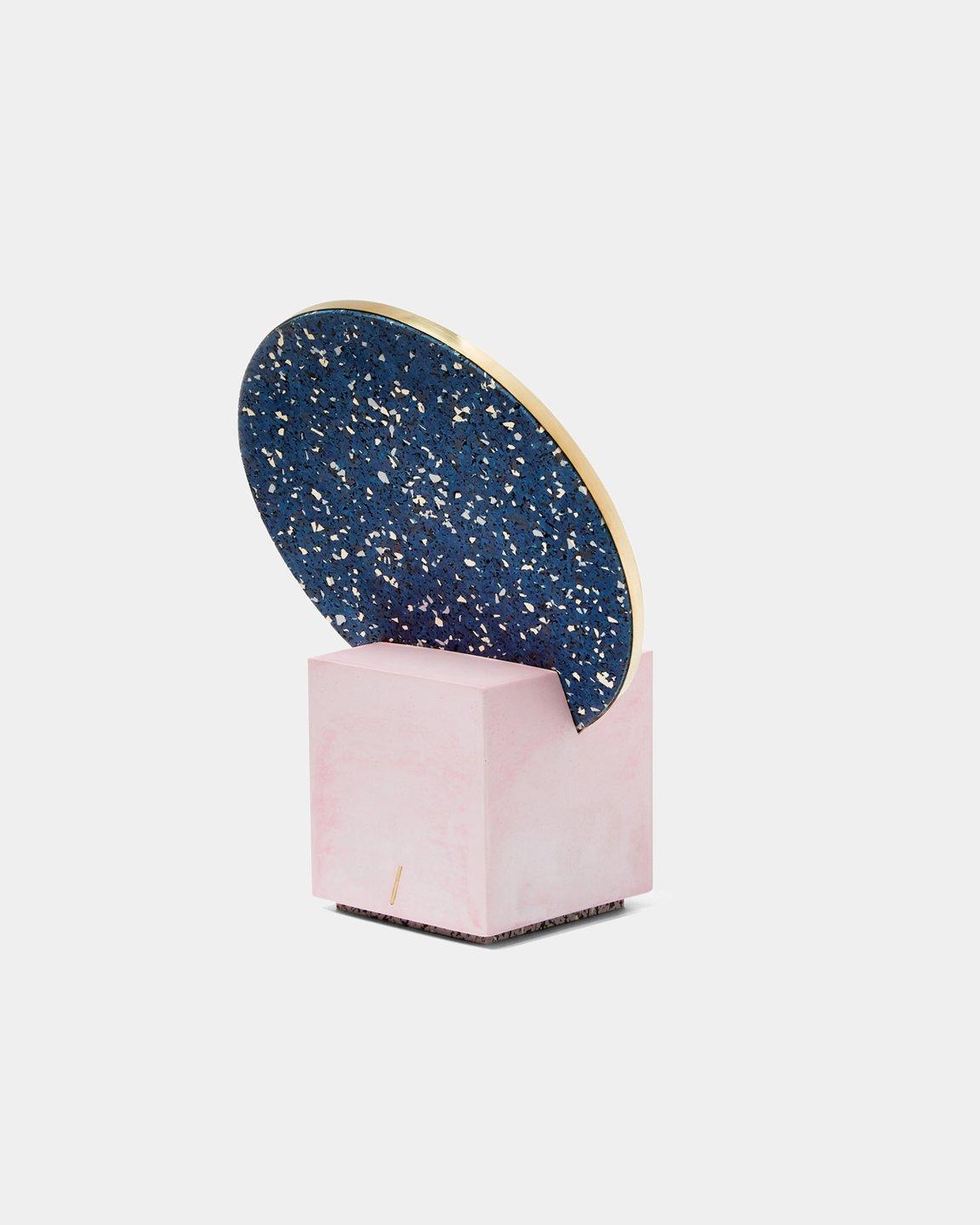 The Vanity Mirror consists of a hand-cast pink cube base with a removable round mirror, edged with a brass frame, and a speckled blue rubber backside. 

Circular mirror with brass edging intersects with the hand-cast cube, showcasing the