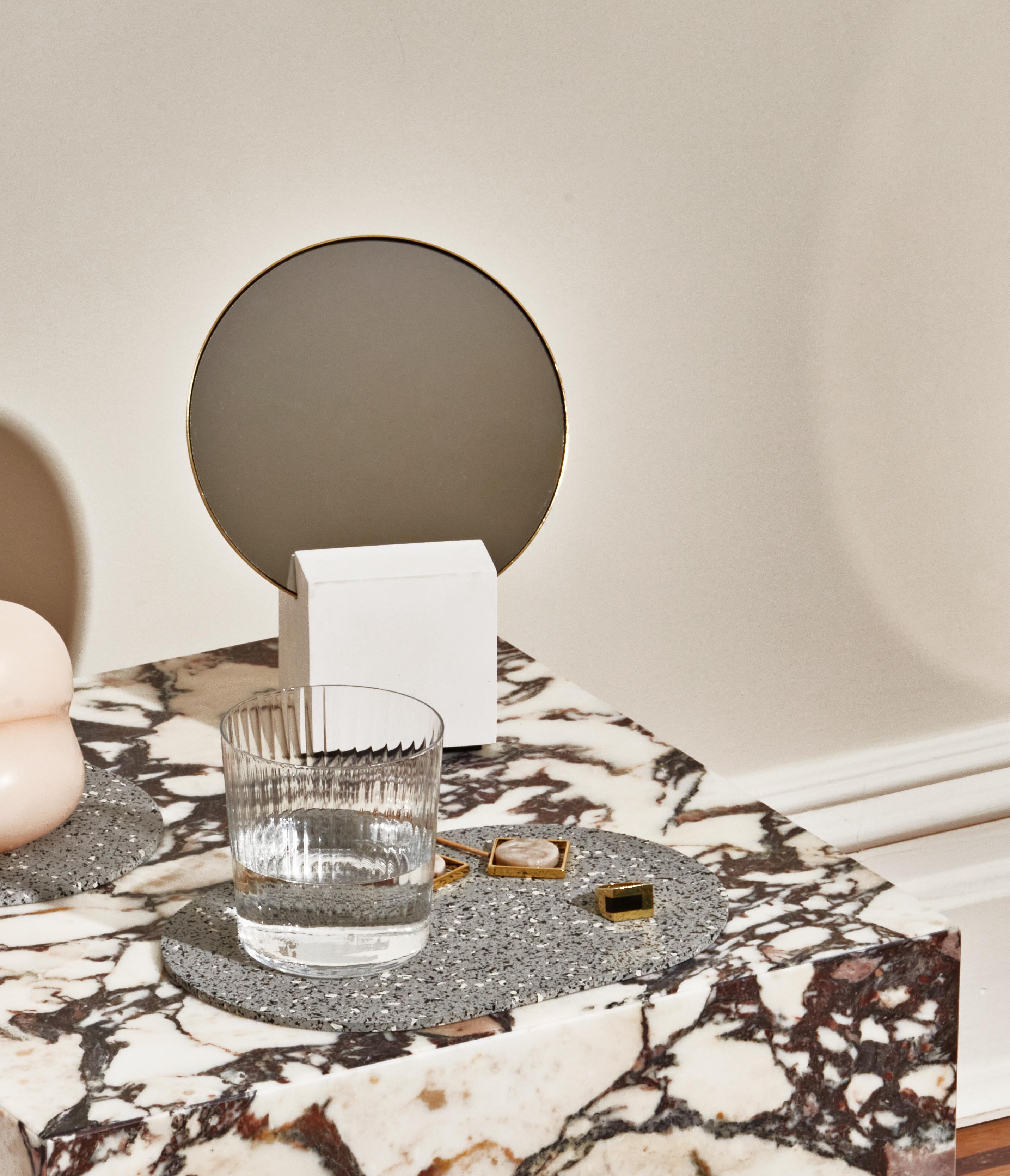 The Vanity mirror consists of a hand-cast white cube base with a removable round mirror, edged with a brass frame. 

Circular mirror with brass edging intersects with the hand-cast cube, showcasing the versatility of its materials. The rubber