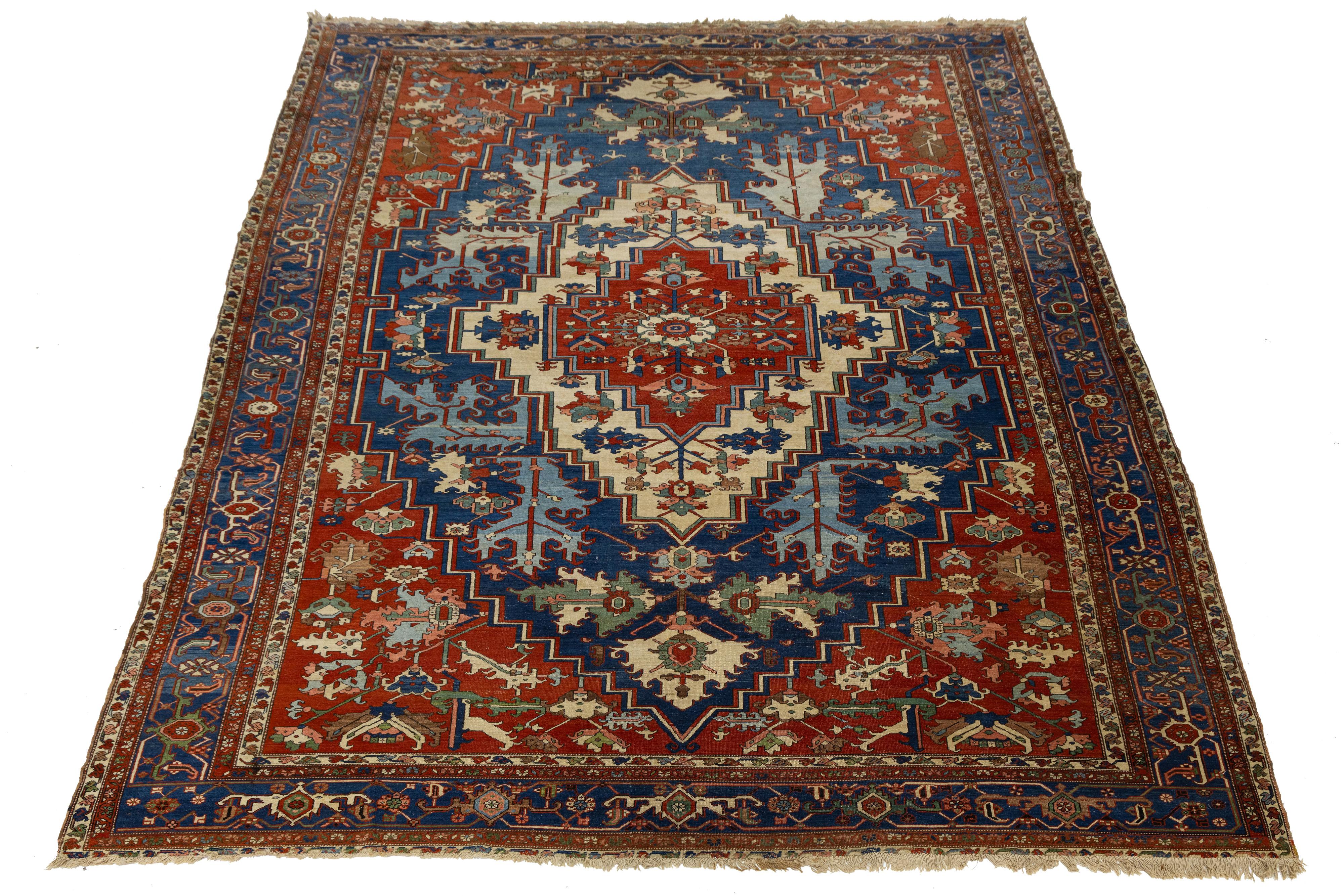 Ce tapis antique Serapi noué à la main présente un médaillon central audacieux et des motifs floraux anguleux dans des tons saturés de rouge, de marine, d'ivoire et de vert. Sa disposition géométrique et son design spacieux reflètent le style