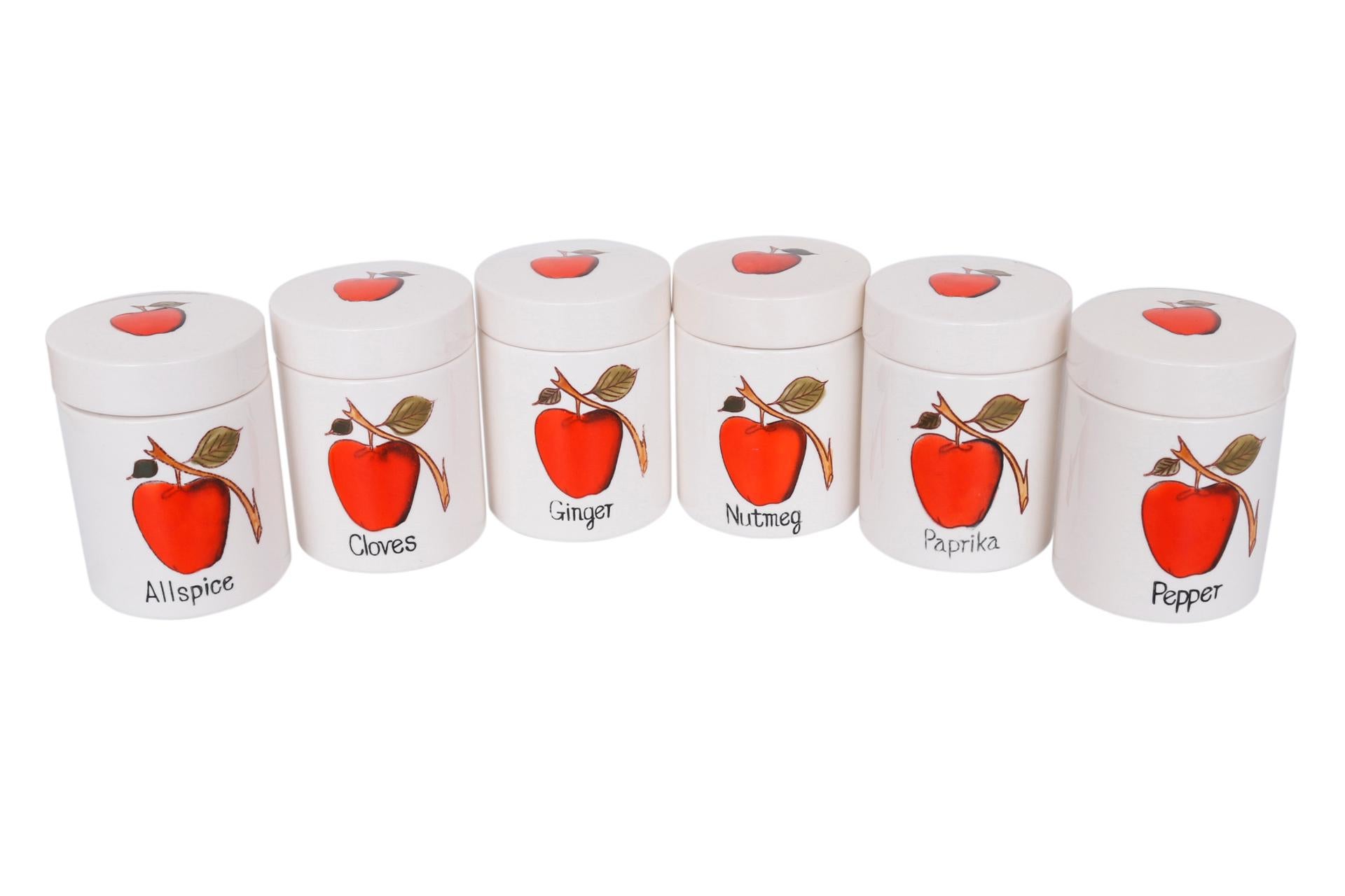 A set of six ceramic spice jars or canisters. Hand painted with red apples in front with small branches on the back. Lids are also decorated with red apples and are lined with a cork rim for an airtight seal. Labeled Allspice, Cloves, Ginger,