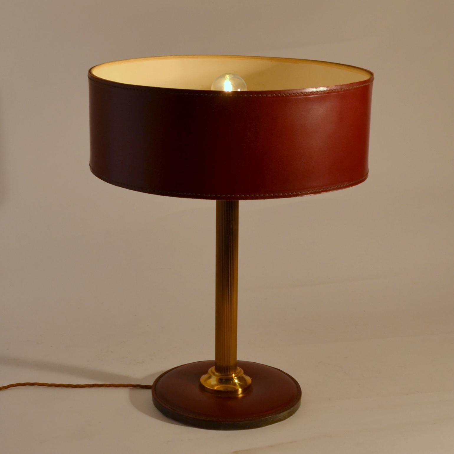 Red brown leather table lamp with a cylinder shade and round base with detailed contrasting stitching standing on a brass stem.
Variations on this model available.