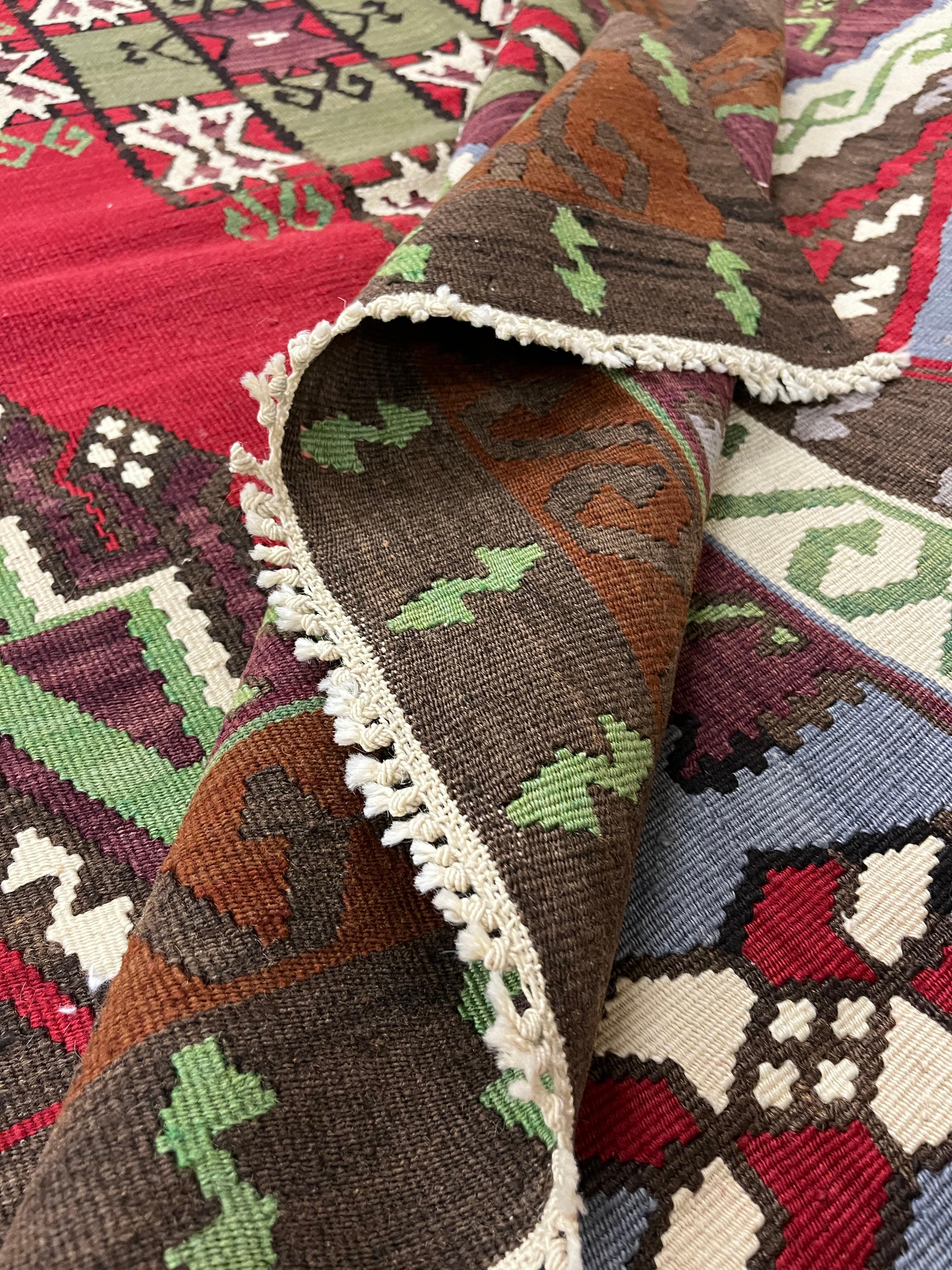 Ce kilim turc en laine fine est un tapis tissé à plat à la main. Il présente un médaillon central audacieux tissé dans des accents de vert, de brun et de crème sur un fond rouge. Il est entouré d'un motif symétrique comportant divers motifs