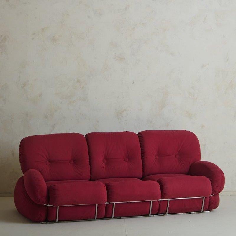 A three-seat 1970s Italian sofa attributed to Adriano Piazzesi. This sofa retains its original red upholstery and features a chrome detail that runs entirely around the sofa. We love the exaggerated proportions and button tufted detailing on this