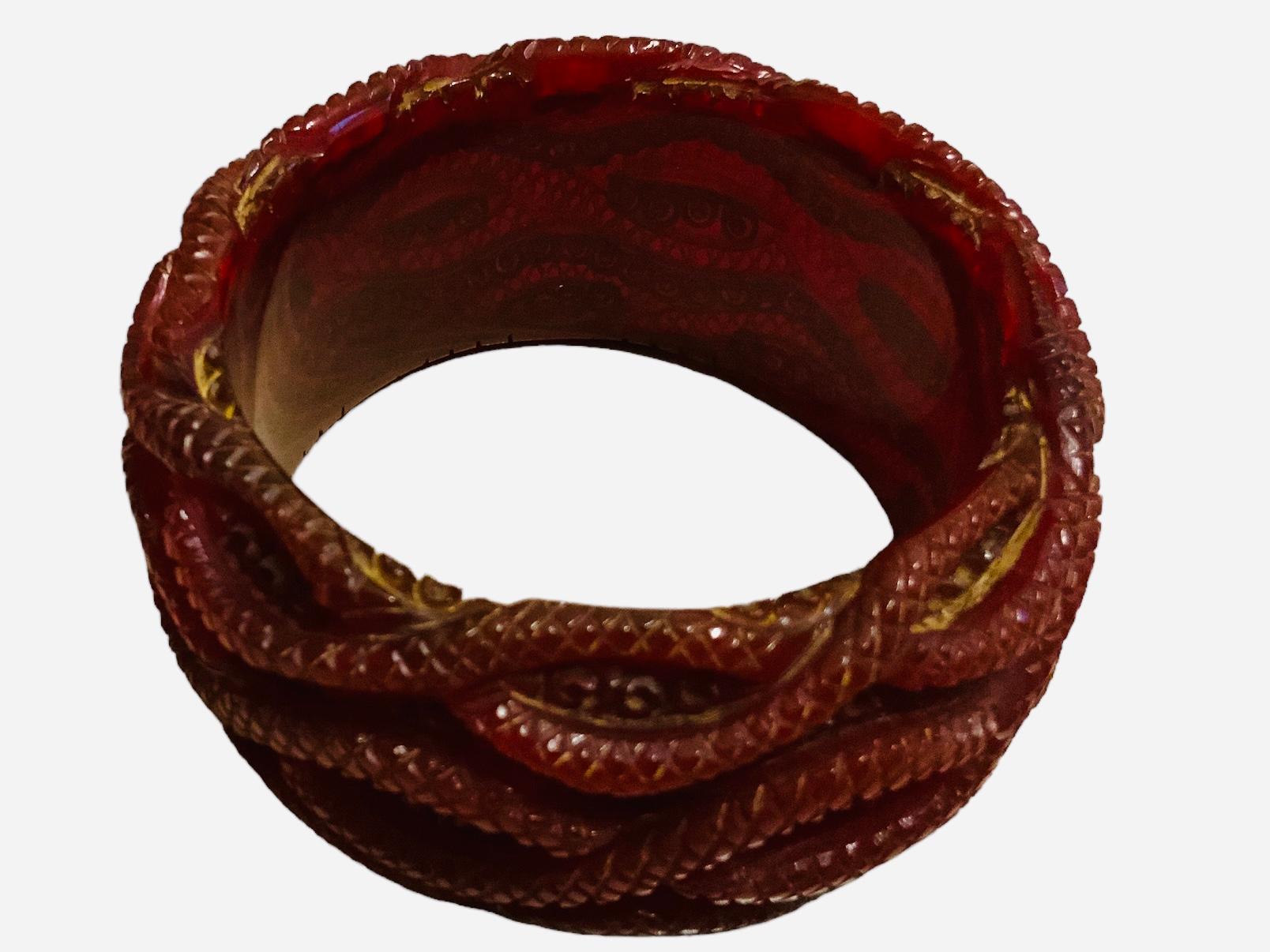 This is a red color carved Bakelite bangle. It depicts a wide bangle that is adorned with a cluster of overlapping snakes like. The “ snakes” are carved with diamonds and rings shaped patterns.