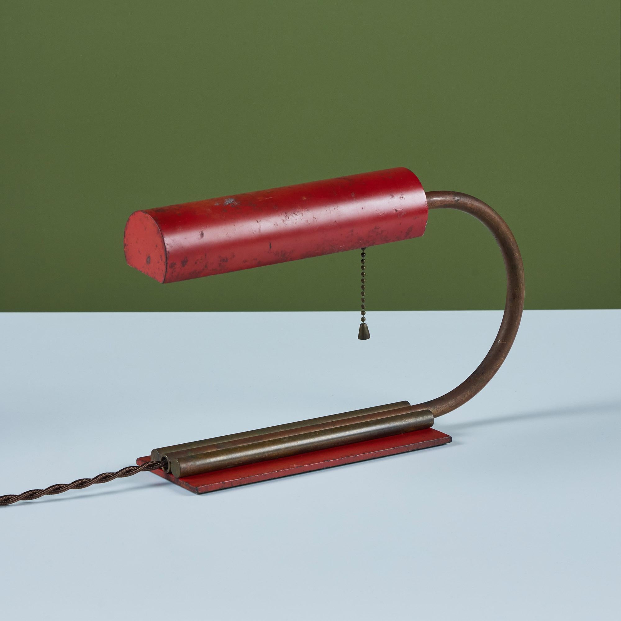 Petite art deco enameled desk or table lamp in the style of Gilbert Rohde, c.1930s. The lamp features a red enameled cylindrical pivoting shade. The brass curved stem of the lamp connects to the rectangular enameled base and is flanked by two brass