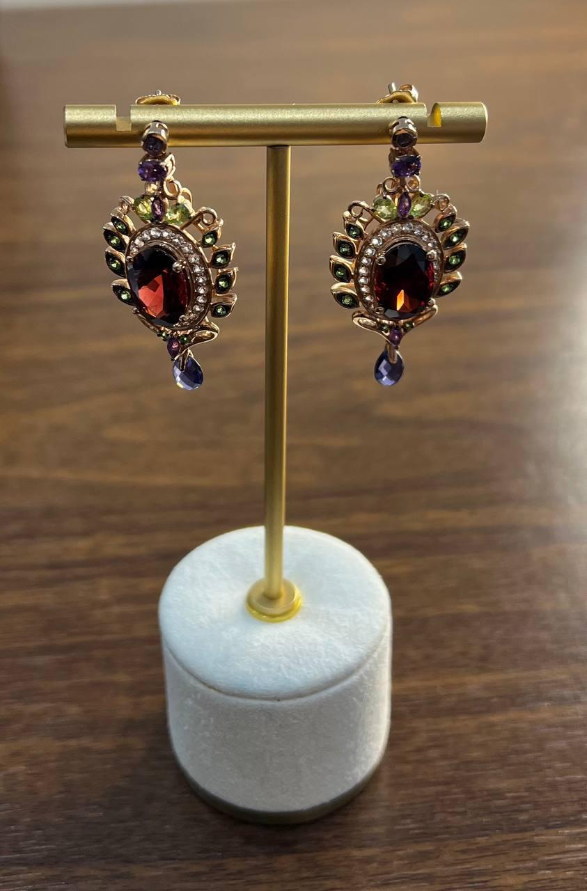 Ces boucles d'oreilles remarquables sont fabriquées en or rose.
Chaque boucle d'oreille est ornée d'un grenat ovale rouge foncé, entouré d'un halo de petites pierres précieuses multicolores étincelantes (saphir, péridot, améthyste), disposées en