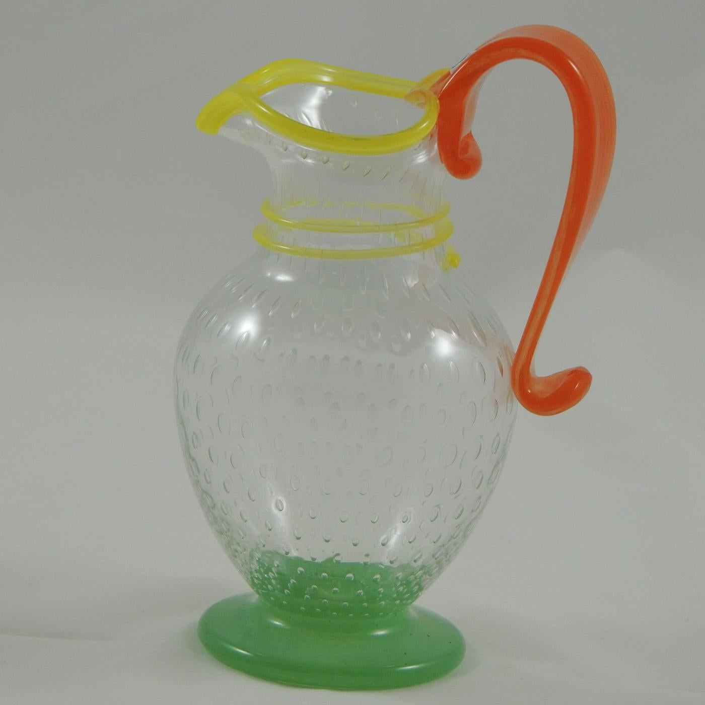 The traditional design of this striking caraffe is made modern by the use of vivid colors. This piece is in mouth-blown Murano glass with a handle embellished by a murrina, typical of the Murano glass art, while the transparent body of the caraffe