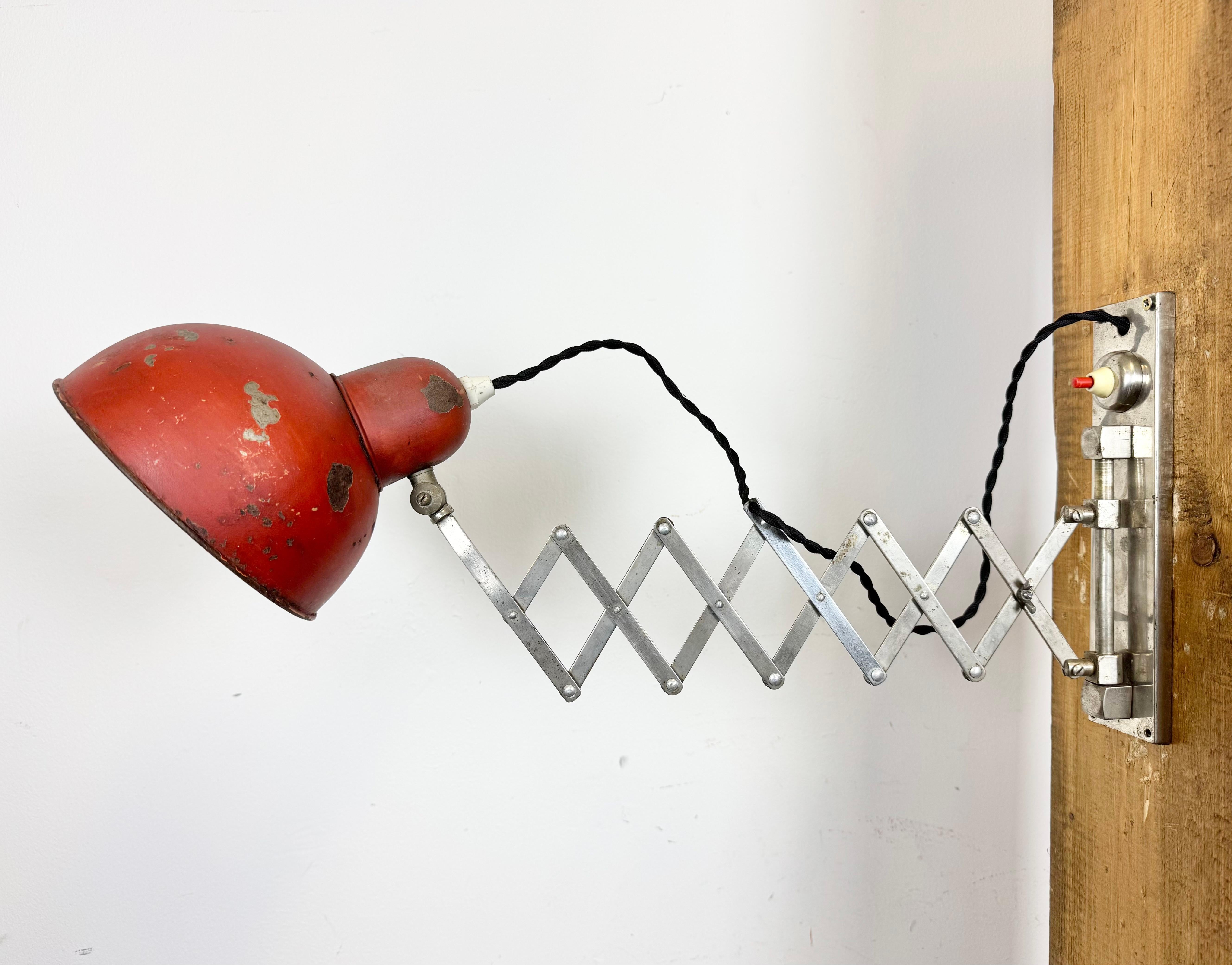 This vintage industrial adjustable scissor wall light was produced in former Czechoslovakia during the 1950s. The lamp has a red metal lampshade. A chrome plated iron scissor arm is extendable and can be turned sideways. .The original socket