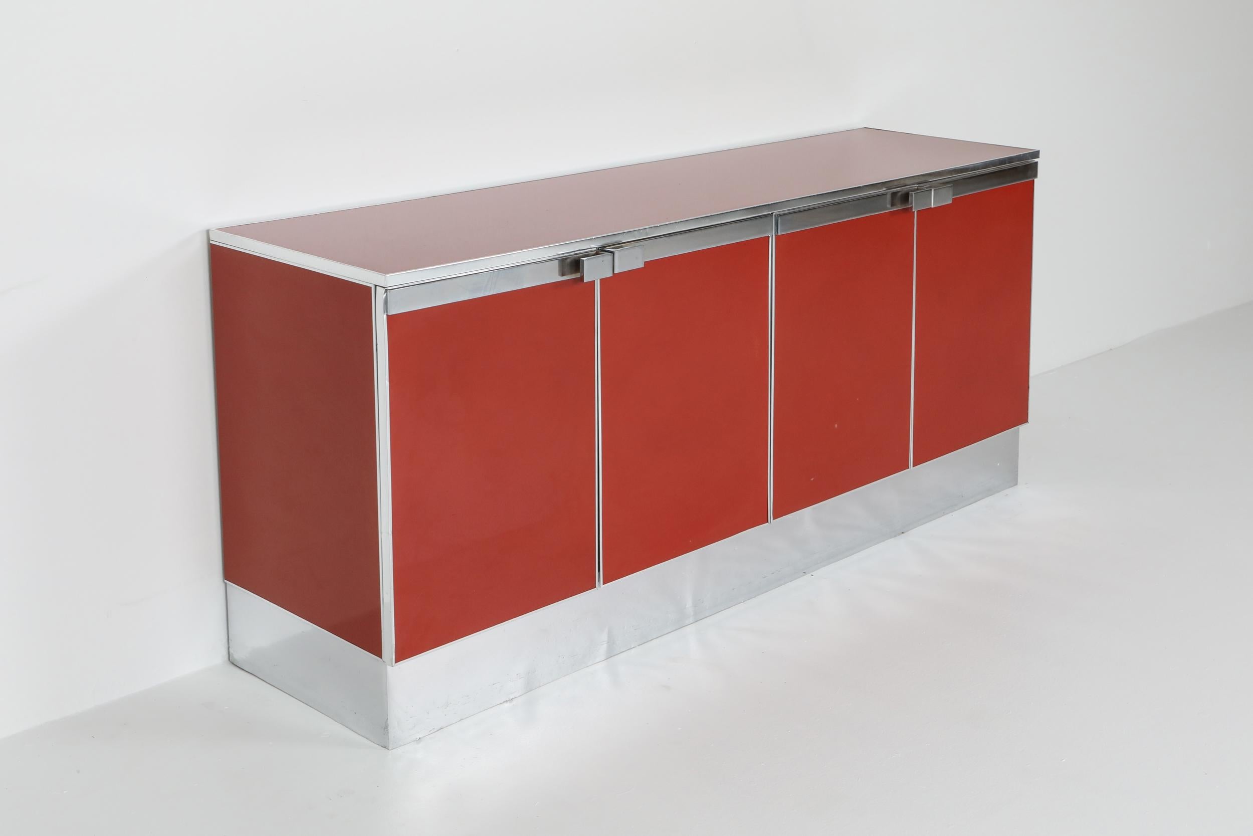 Eclectic Postmodern sideboard in red lacquer with chrome detailing all around.
The credenza has four lacquered doors that hide shelving and a drawer.
Would fit well in a metropolitan chic decor inspired by Romeo Rega, Willy Rizzo, and Mario