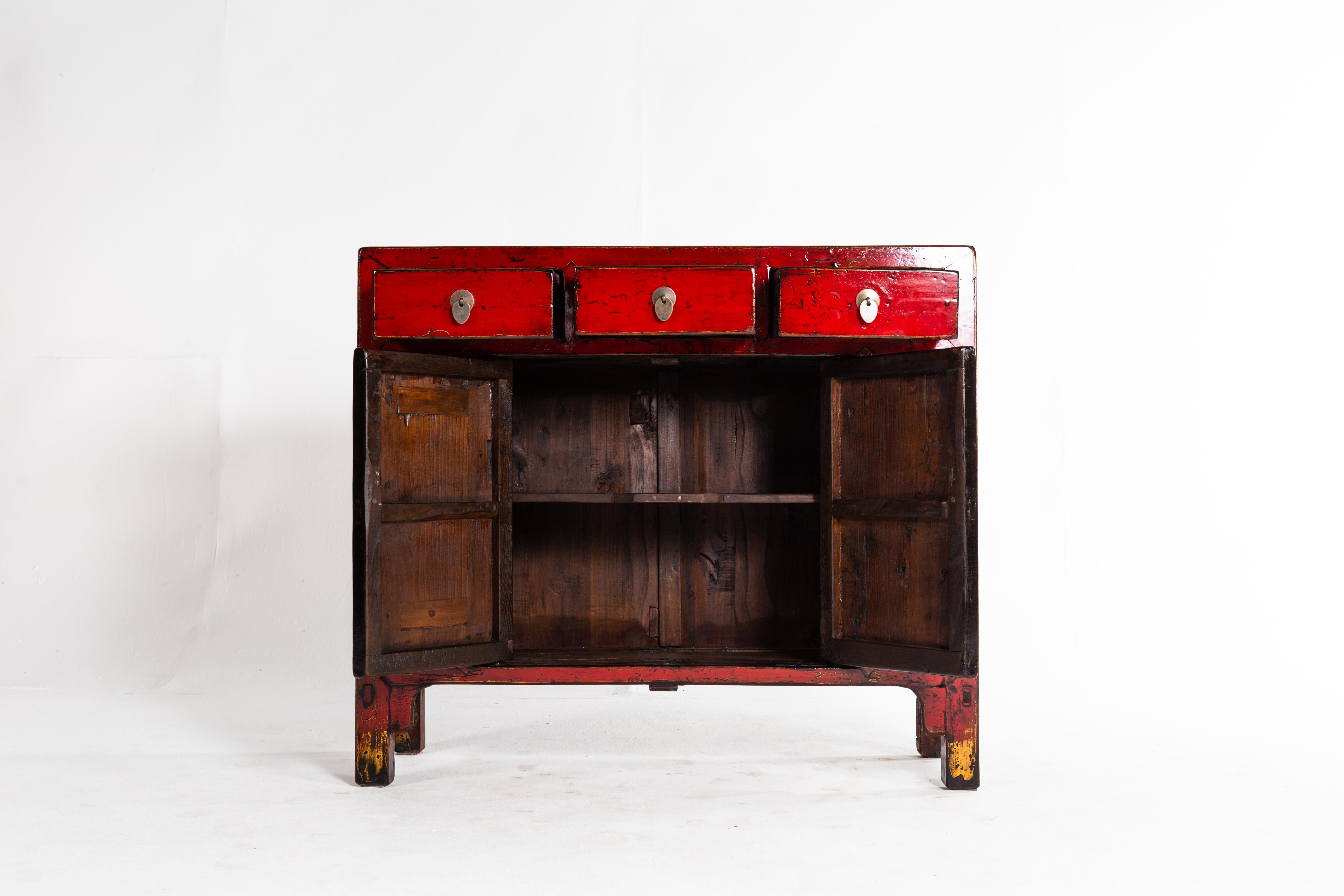 This cabinet is from Shandong, China and was made from elm, pine, and lacquer, circa 1920. The piece features 3 drawers, a pair of doors, red lacquer, and a shelf for storage. Wear consistent with age and use.