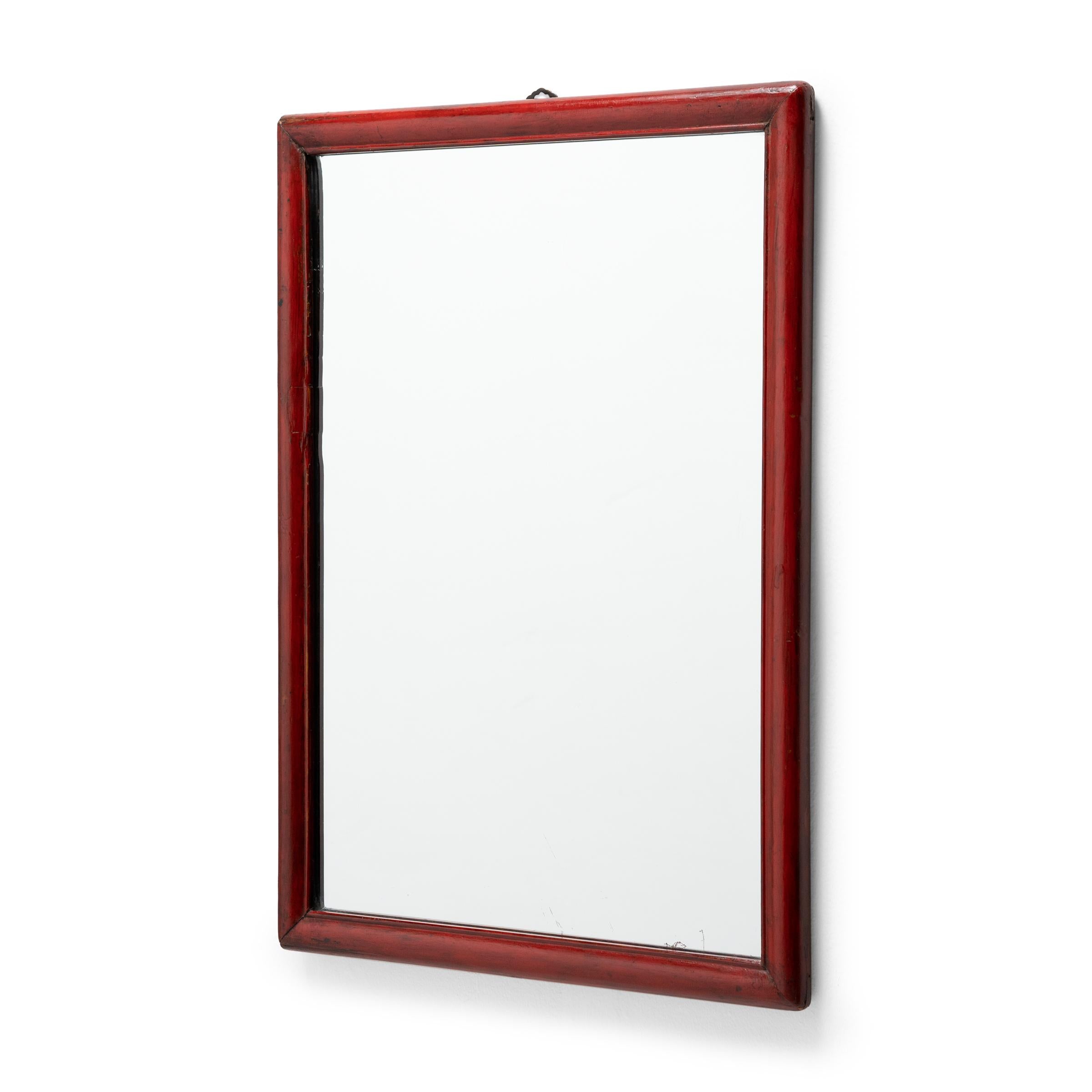 This rectangular wall mirror features a simple wooden frame with a beaded trim and a rich, red lacquer finish. Rounded corners on one side and sharp corners on the other suggest this mirror was originally part of a mirrored table screen, placed