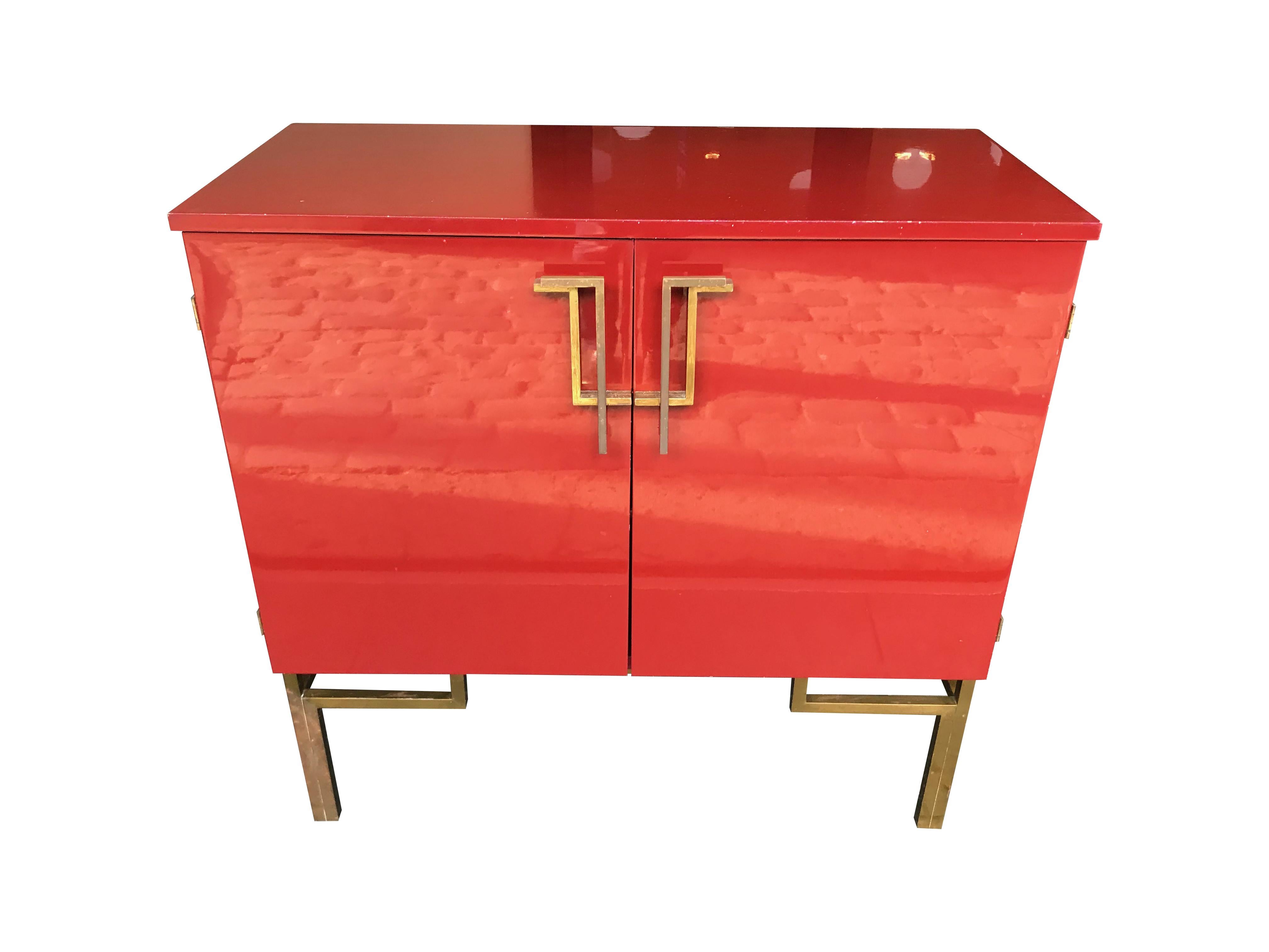 A red lacquered 1970s bar cabinet with brass chinoiserie detailing. The top opens to reveal a recessed mirrored serving area. Two doors on the front have geometric brass handles that open to reveal shelves for glasses and bottles with mirror base