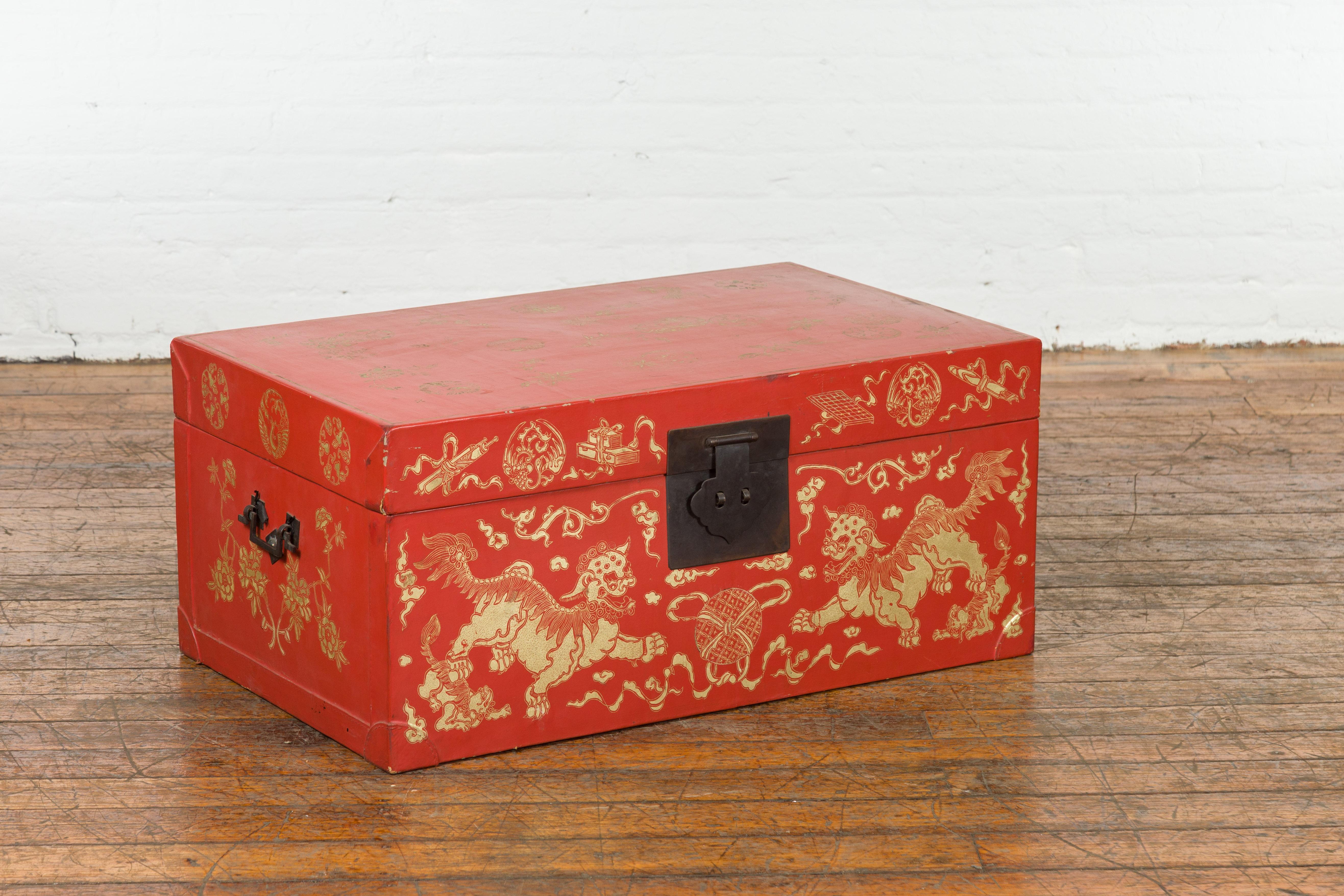 A Chinese red lacquered blanket chest from the 20th century, with gilt décor depicting guardian lions, flowers, scrolling clouds and scholar objects, as well as yellow fabric lining and iron hardware. Created in China during the 20th century, this