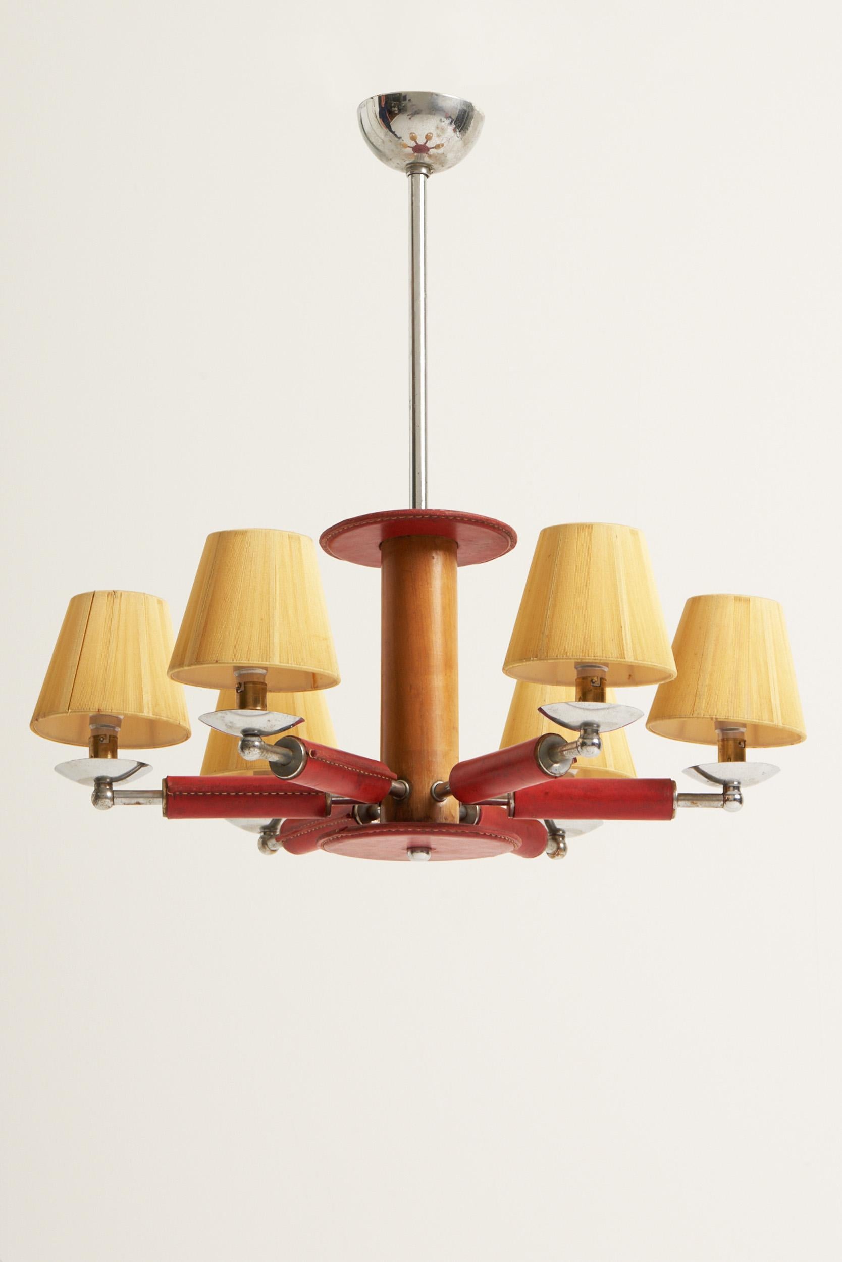 A saddle stitched red leather, oak and nickel six-armed ceiling light by Jacques Adnet (1900-1984). With its original shades. 
France, Circa 1955.