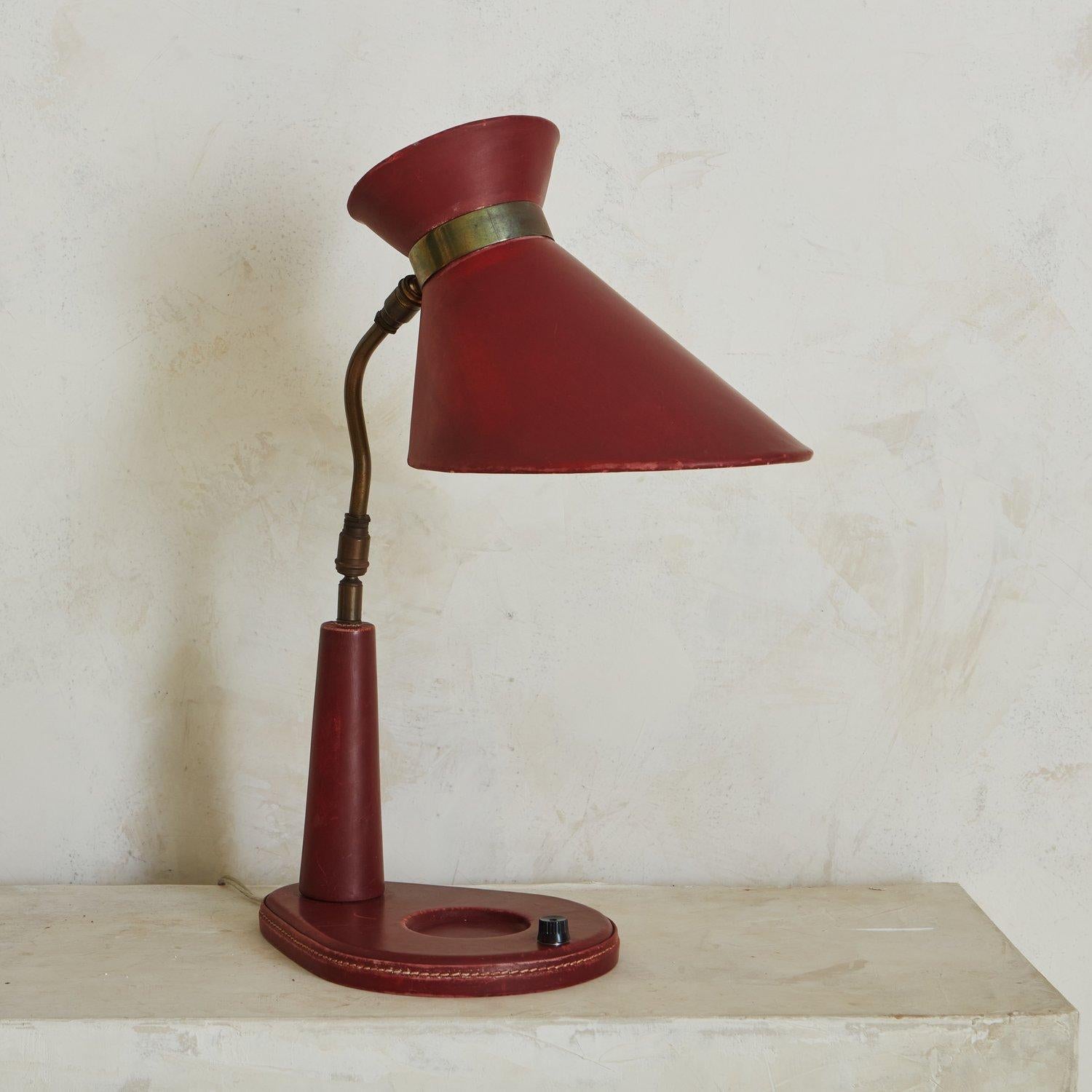 A 1950s desk lamp in the style of Jacques Adnet. This lamp has a shade and body clad in red leather with stitch detailing. It has a curved, adjustable brass stem with a beautiful patina. This lamp has a circular catchall detail on the base next to a