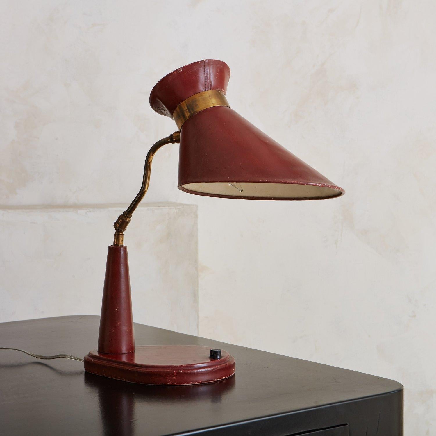 A 1950s desk lamp in the style of Jacques Adnet. This lamp has a sculptural shade and body clad in red leather with a black acrylic switch. It has a curved, adjustable brass stem with a beautiful patina. Sourced in France, 1950s.