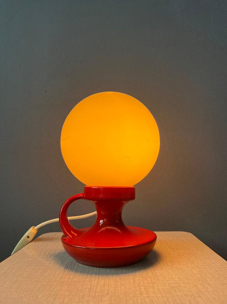 Petite lampe de table en céramique rouge d'Allemagne de l'Ouest avec abat-jour en verre opalin. La lampe nécessite une ampoule E14 et dispose actuellement d'une fiche de connexion à l'UE.

Informations complémentaires :
MATERIAL : Céramique,