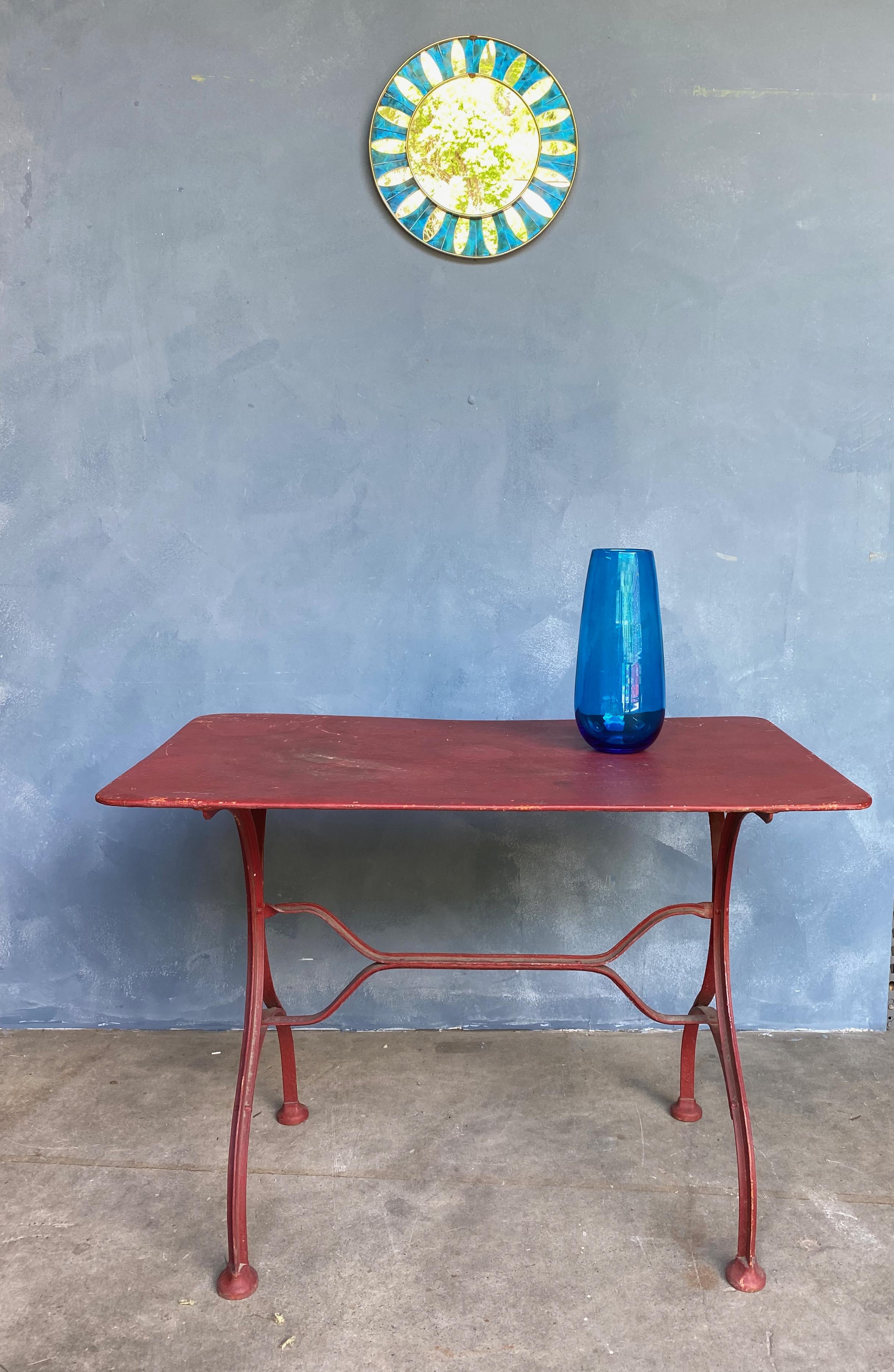 This red painted French iron garden table will add a pop of color and vintage charm to any outdoor space. The classic French bistro style of the iron base and the solid metal top are a nod to the industrial design of 1920s France. Painted in a