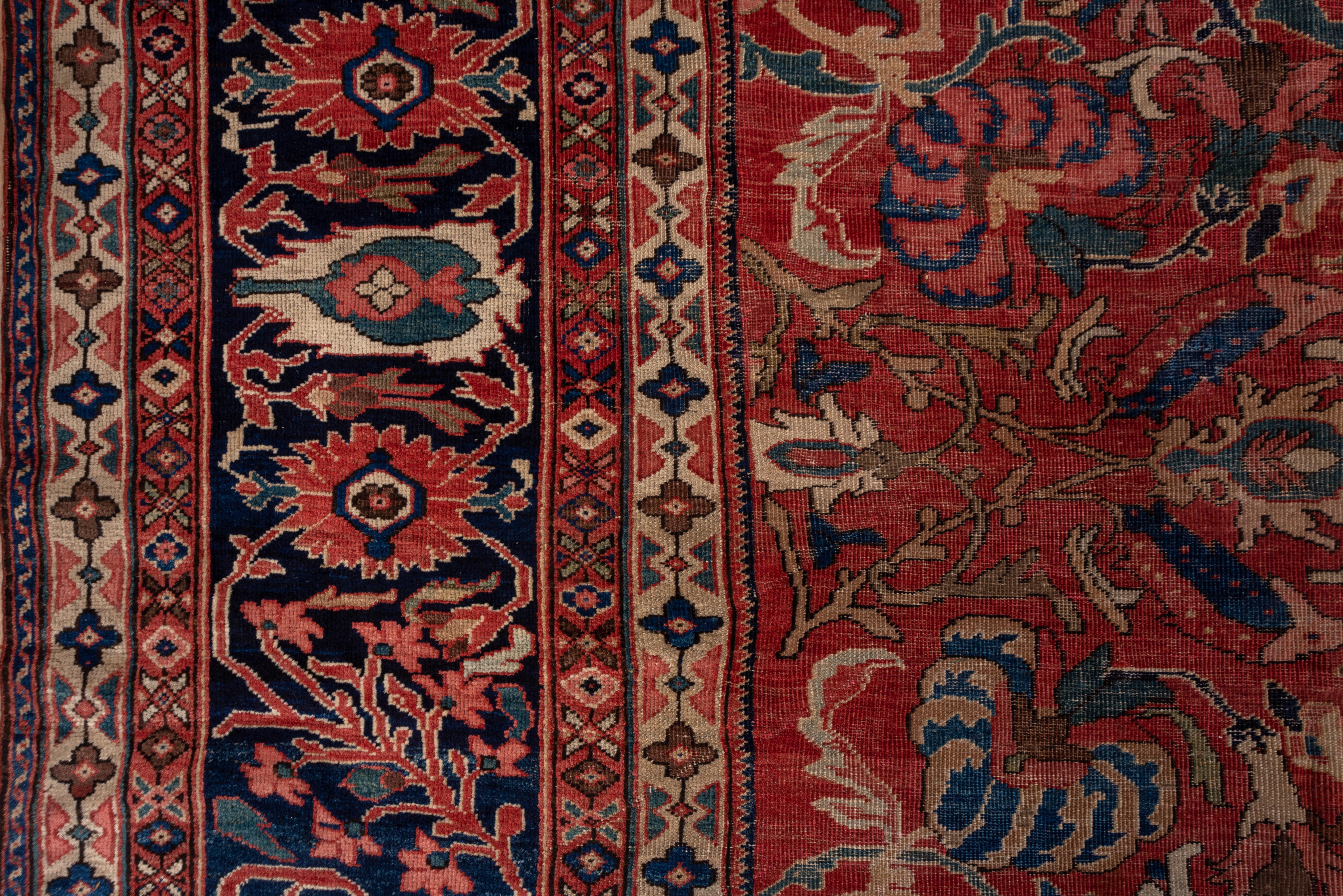 This magnificent west Persian village carpet with a splendid natural dye palette features a tomato madder field centered by a royal blue rosette and displaying teal doubly curved leaves, petal rosettes, striped palmettes and vinery segments. Royal