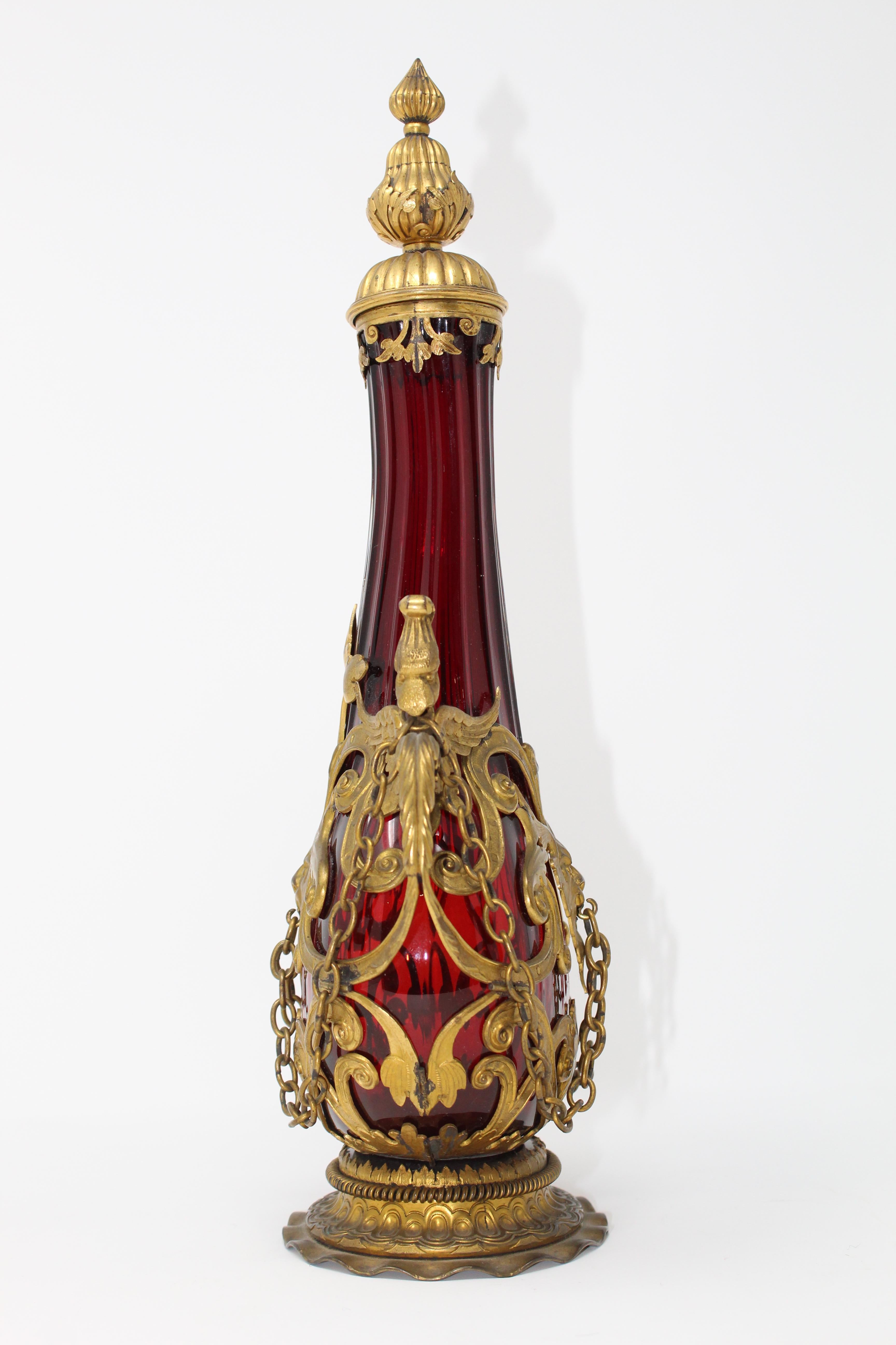 A red ribbed-glass gilt-metal mounted flask.
The metal mounted onto the bottle fits around it perfectly and is shaped and held into place by little hinges. The bottle features 'handles' in the shape of griffin heads with wings spanning under them.