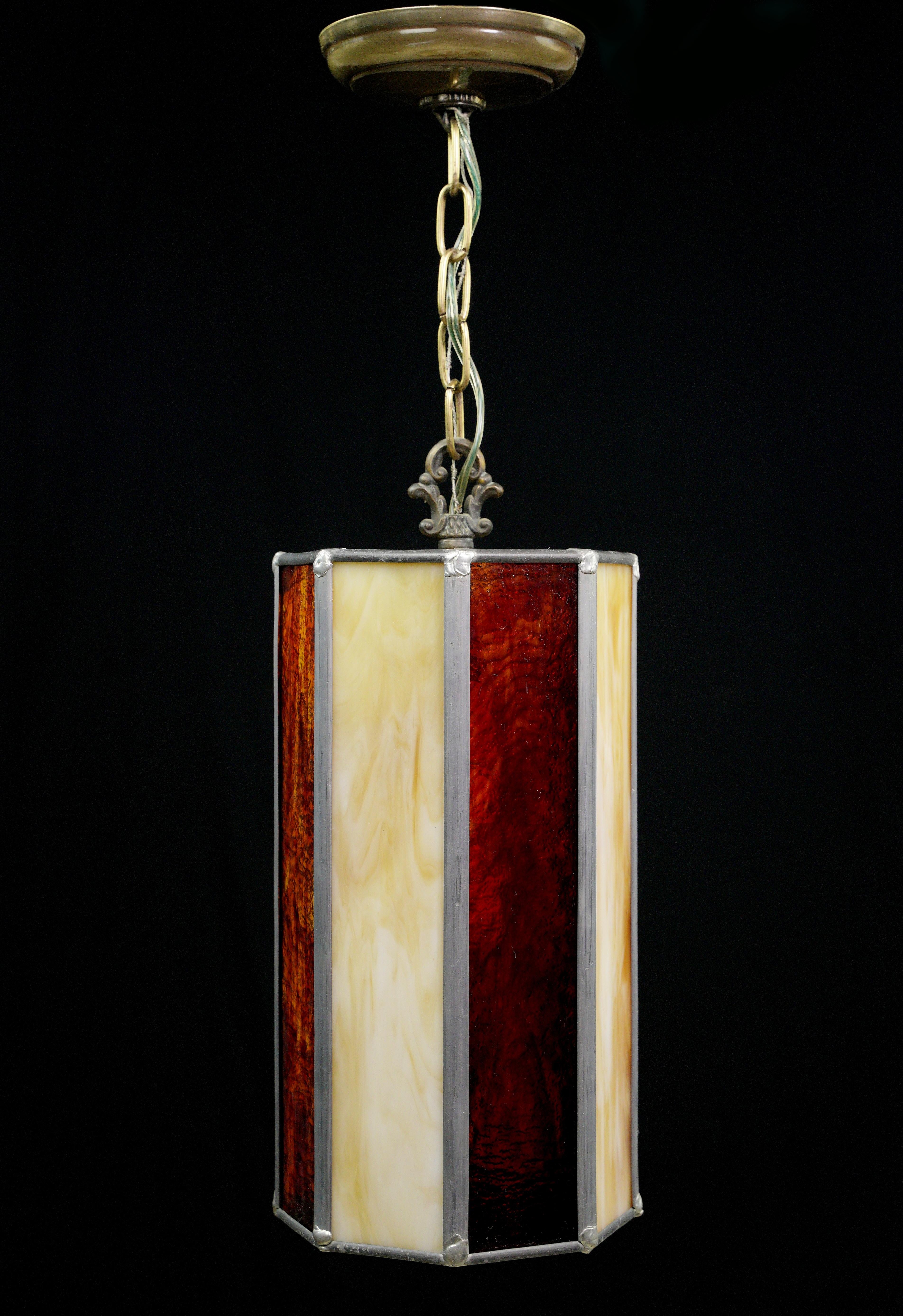 This brass chain pendant light is a captivating piece, fusing artistry and functionality. The leaded stained glass in red and tan hues casts a mesmerizing glow, while the brass chain adds a touch of vintage charm, elevating any interior with its