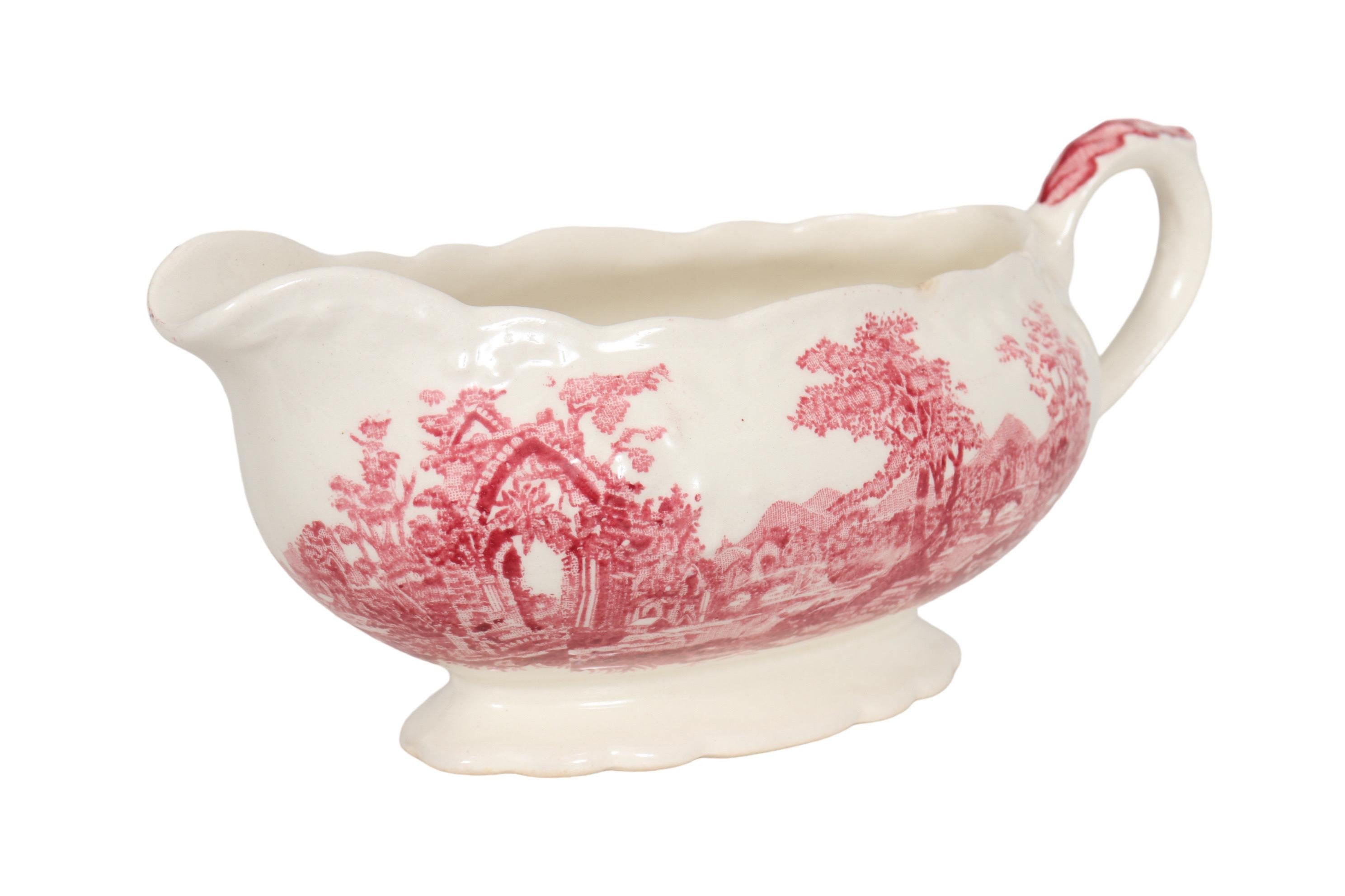 A transferware ceramic gravy boat with a scalloped edge, made by Taylor, Smith & Taylor. Decorated in their red English Abbey pattern. The pattern shows an abbey and trees, beside a river with a bridge. Maker's mark visible underneath.