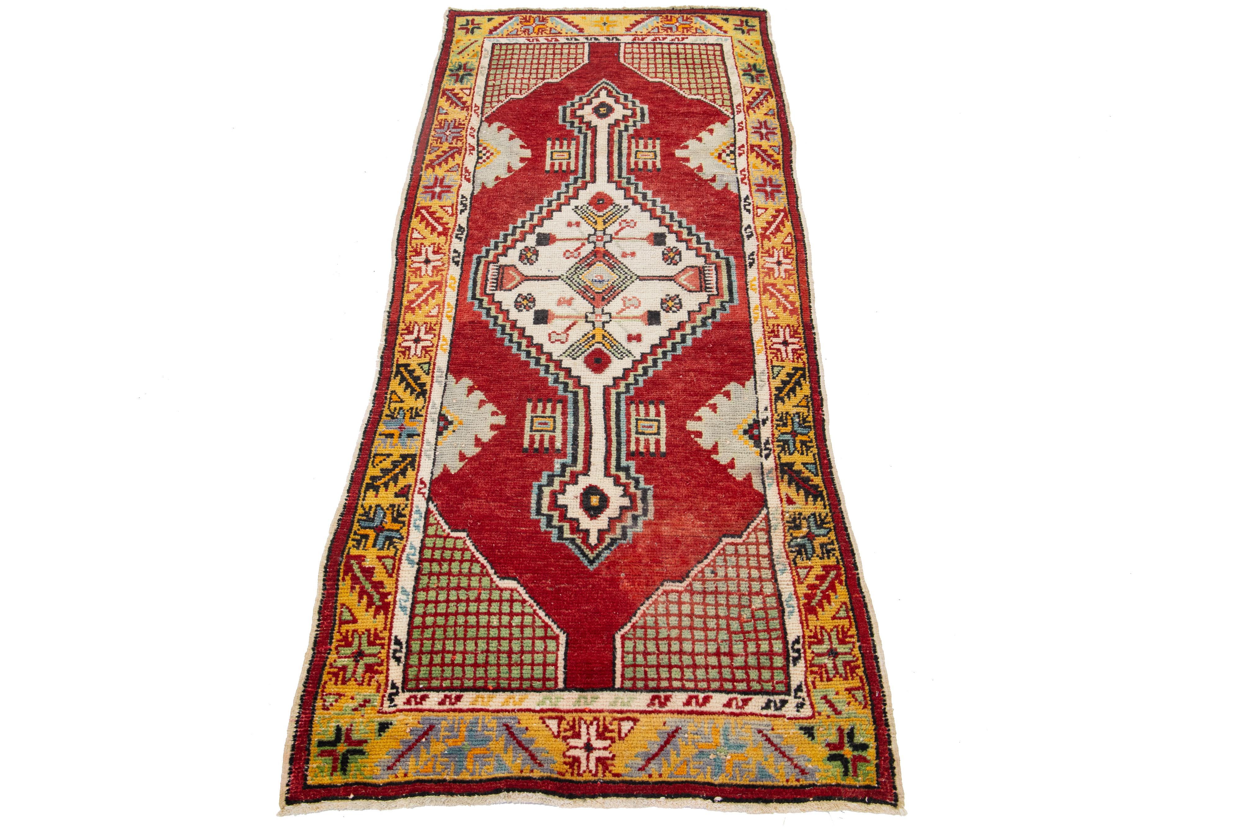 A hand-knotted vintage Anatolian rug features a geometric design on a red field, with ivory, green, and yellow accents throughout the piece.

This rug measures 2'7