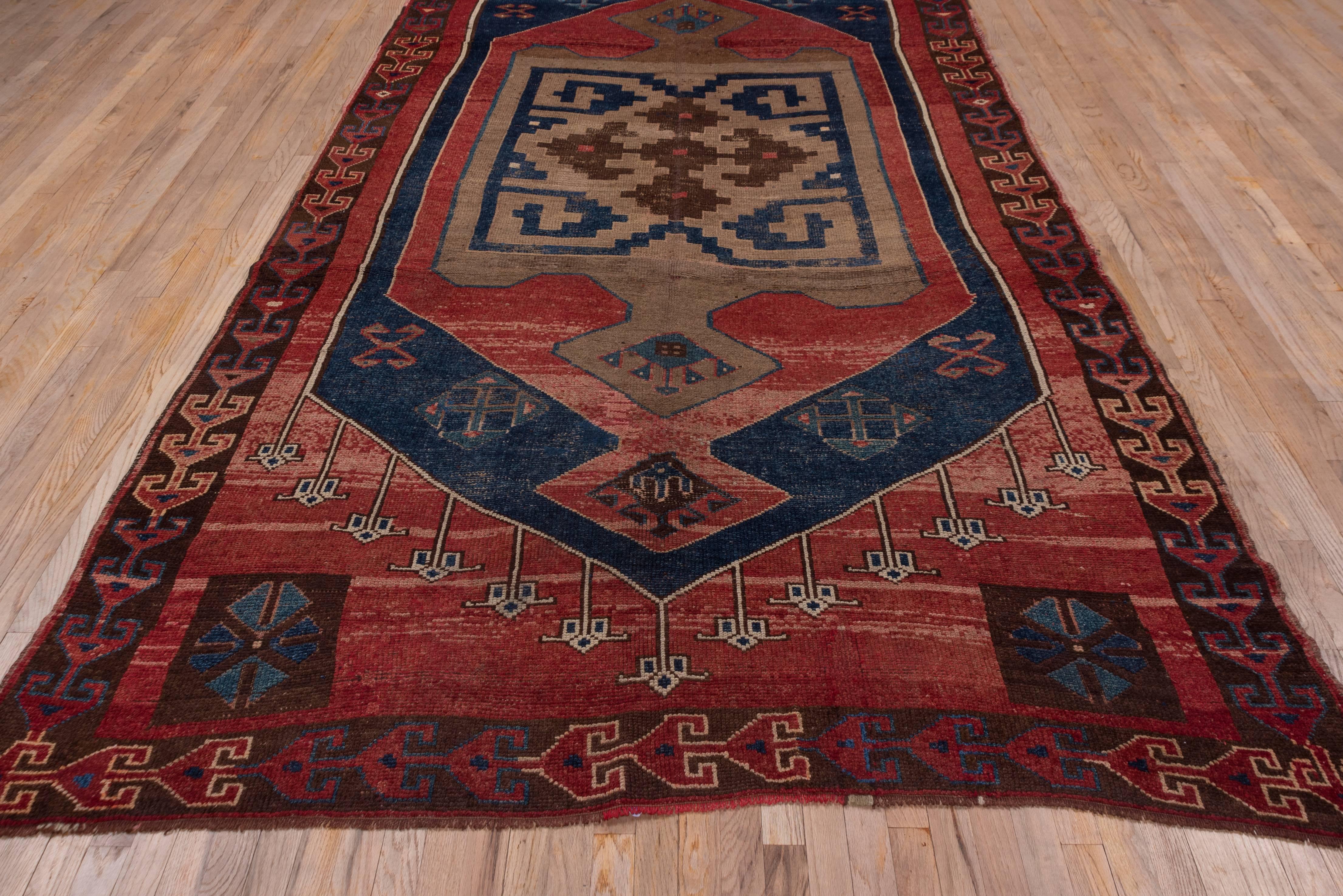 This boldly geometric long carpet features a red field with dark blue hexagonal subfield enclosing a pendant oblong red medallion and an olive inner medallion with an enormous reciprocal pattern in red and dark blue. The dark brown single border
