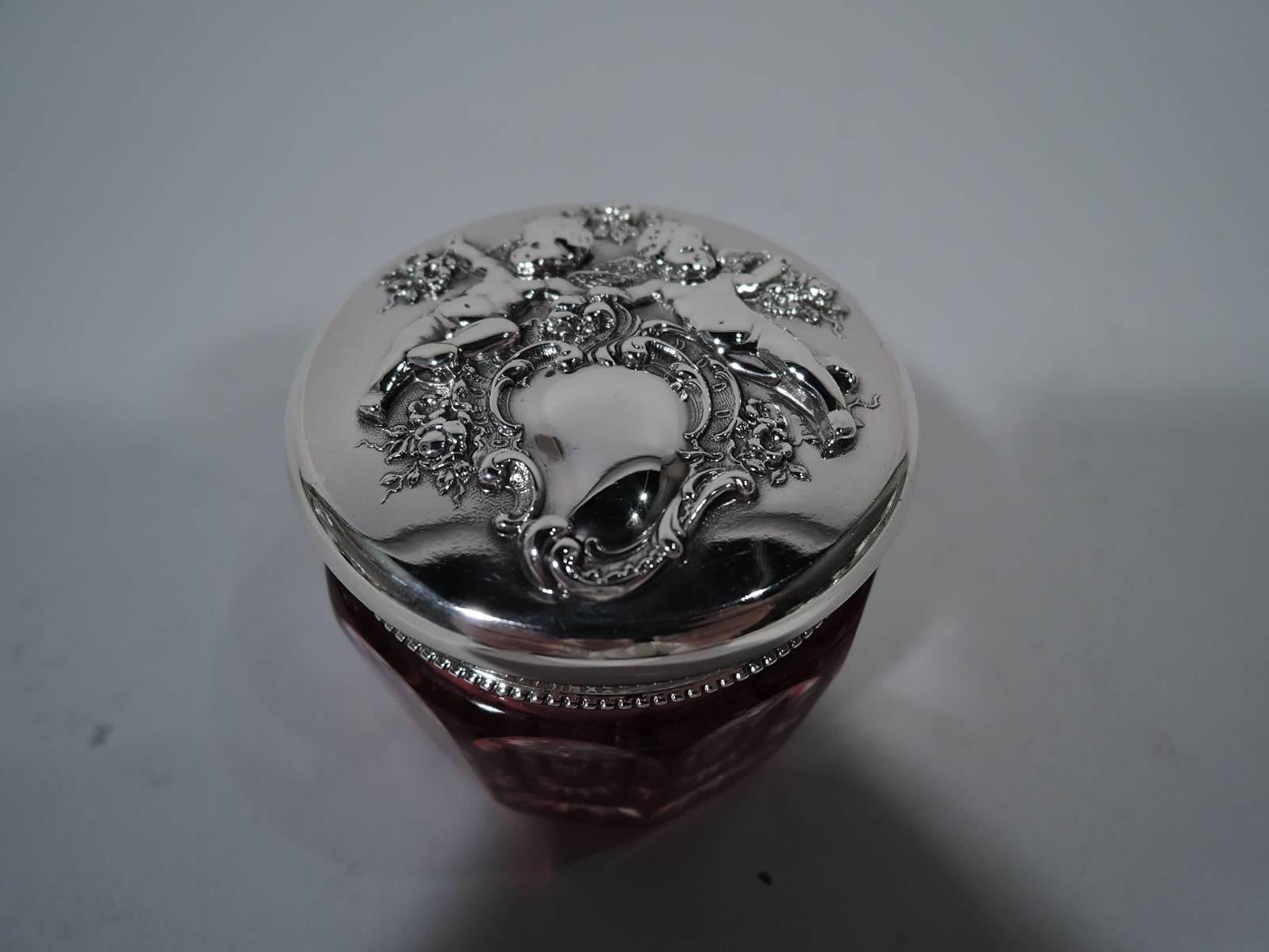 Art Nouveau sterling silver and glass vanity jar. Made by Reed & Barton in Taunton, Mass. Faceted clear glass jar with cranberry flashing. Sterling silver cover with dentil rim and chased and repousse cherubs gripping bouquets and reclining on