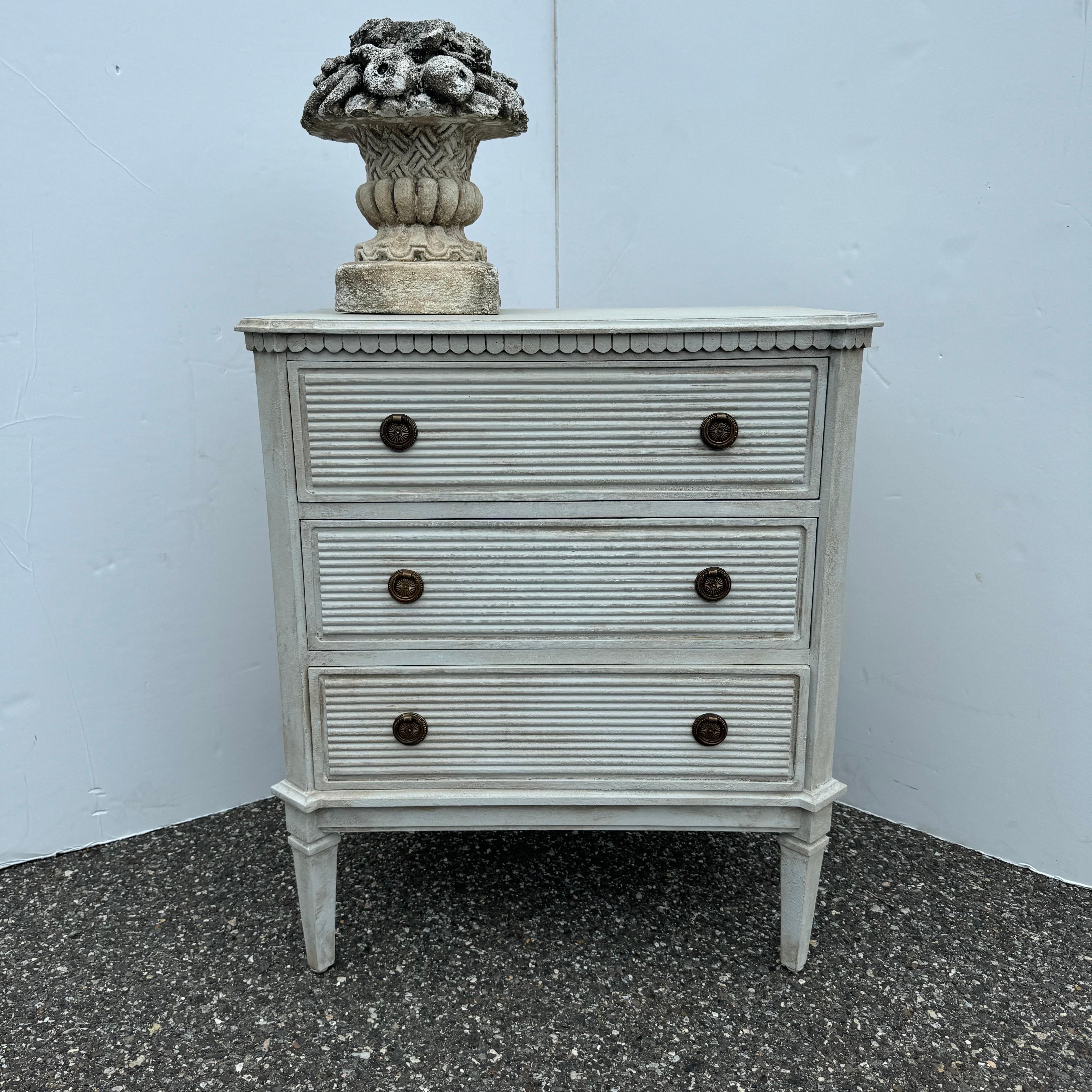 Cassettiera in stile gustaviano dipinta a mano, di colore bianco/grigio, realizzata in legno massiccio con finitura anticata applicata a mano e ferramenta ad anello in ottone. Queste classiche commodes a tre cassetti in stile svedese, con bordatura