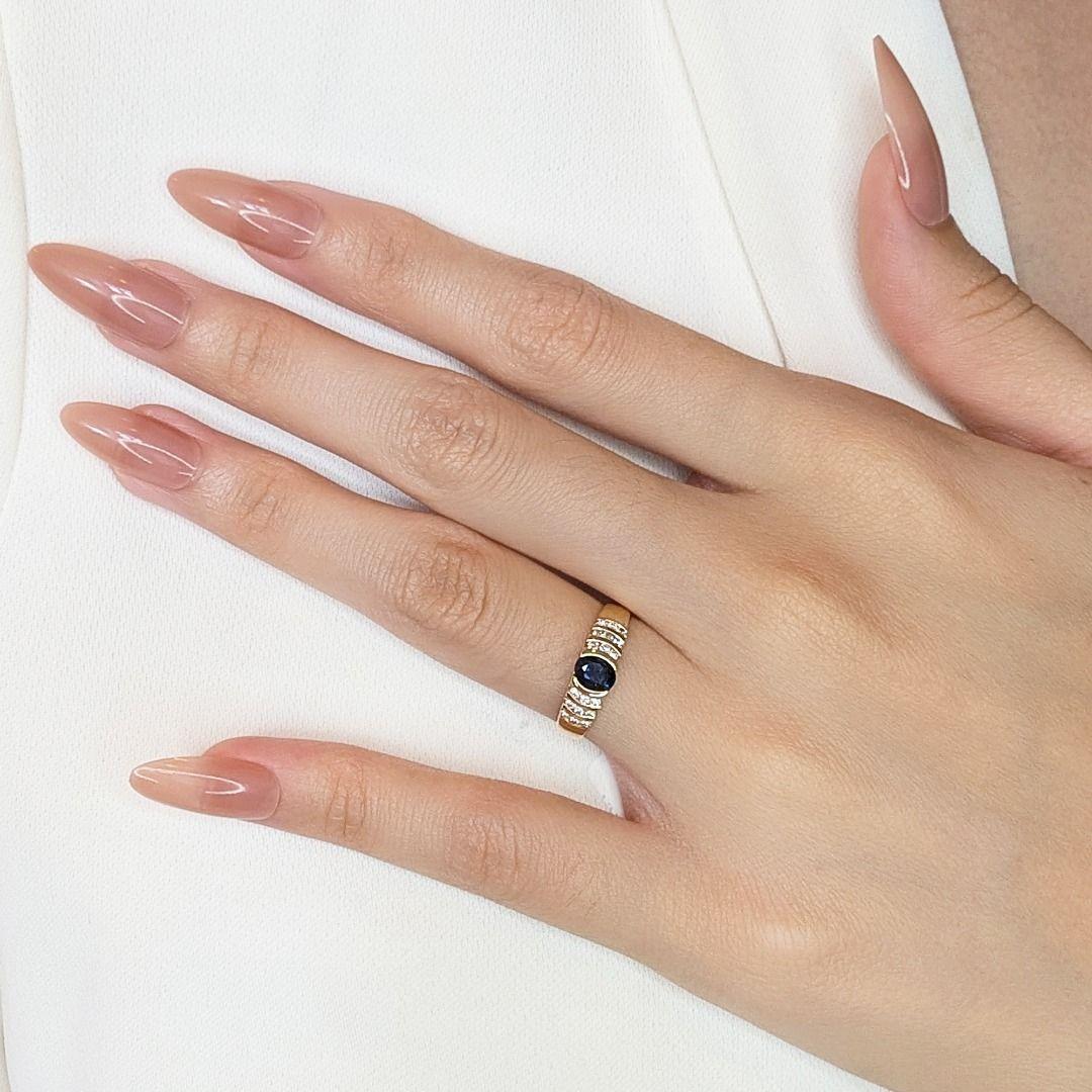Este precioso anillo de oro amarillo de 18 quilates contiene un cautivador zafiro ovalado de 0,2 quilates, de un profundo tono azul oscuro. Acentuado por 18 diamantes brillantes redondos naturales que suman 0,09 ct (color H, claridad VS-SI), este