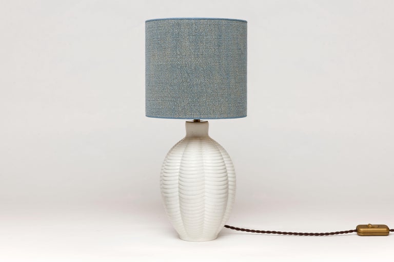 Lovely small refined white ceramic table lamp with serene pattern by Upsala Ekeby, Sweden, 1960.
The shown shade is included. It's handmade, executed from a high end blue fabric with a golden interior.

Upsala Ekeby was, next to Gustavsberg and
