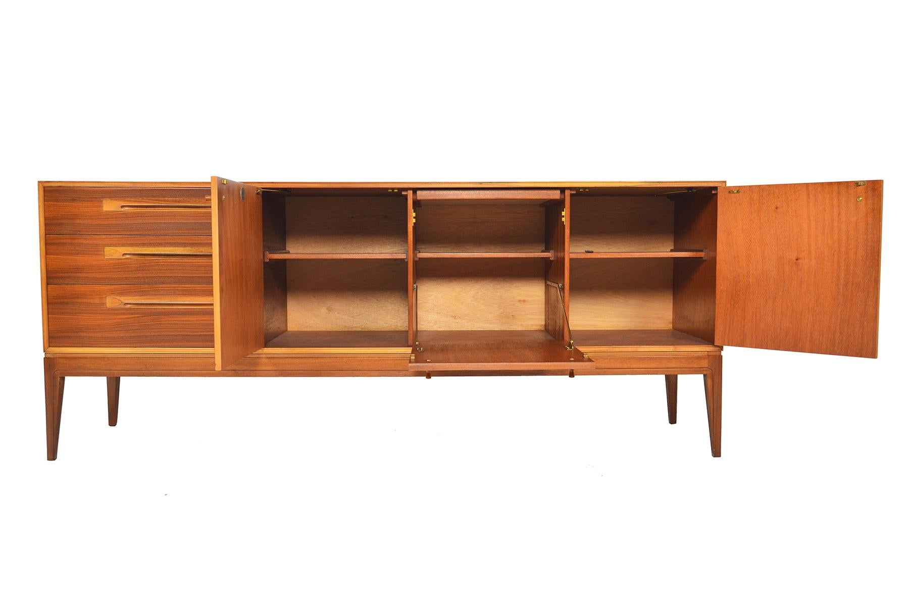 This stately midcentury credenza was crafted by A.H. McIntosh in the late 1960s. Beautiful, multi-tonal Tola wood lines the doors and case. A bank of three drawers sits to the left of three bays outfitted with fixed shelves. A center bay offers a