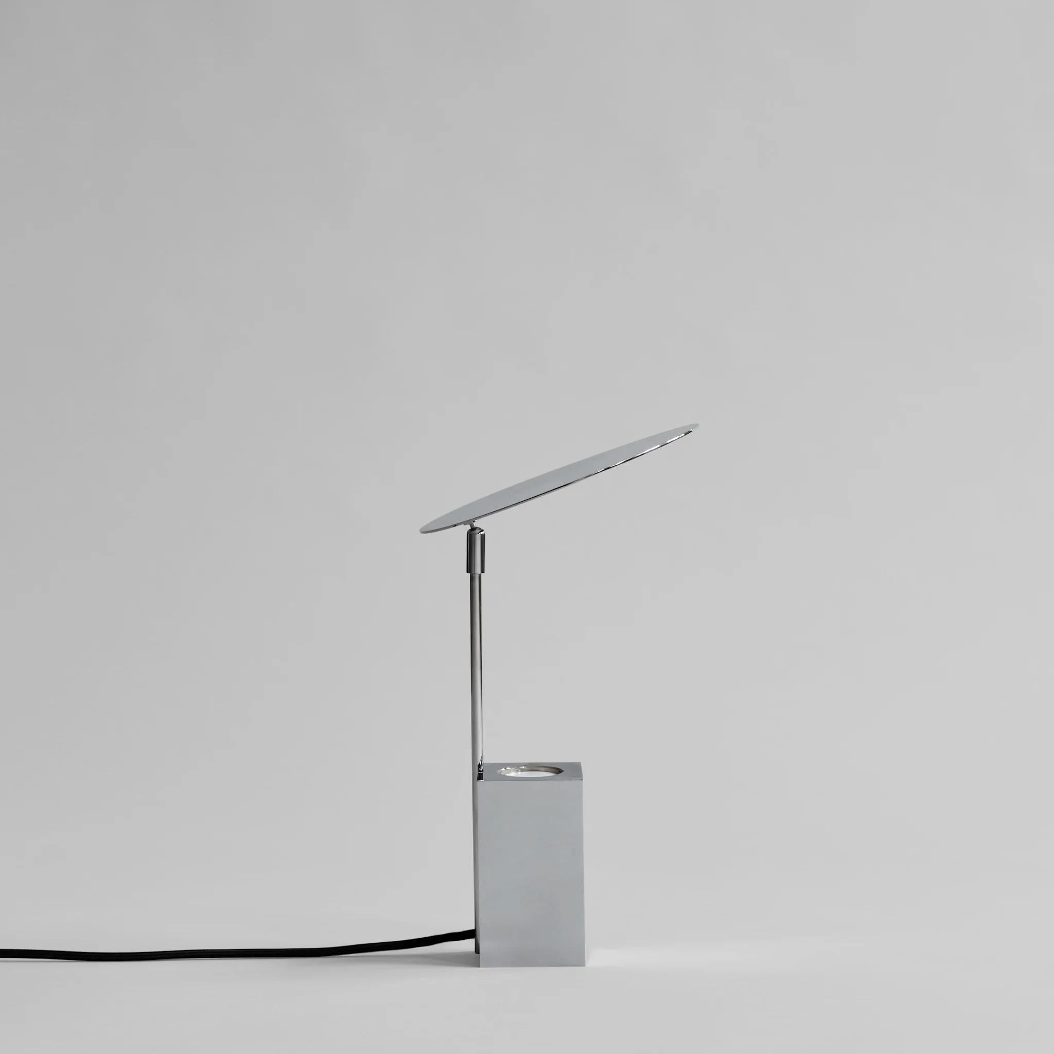 Reflect table lamp in contemporary chrome-plated metal.
 
The lamp's elegant silhouette is made up of a slim, rectangular metal cage with a mirror chrome finish, which houses a concealed E14 socket. 
A white opal glass cover sits above the socket,