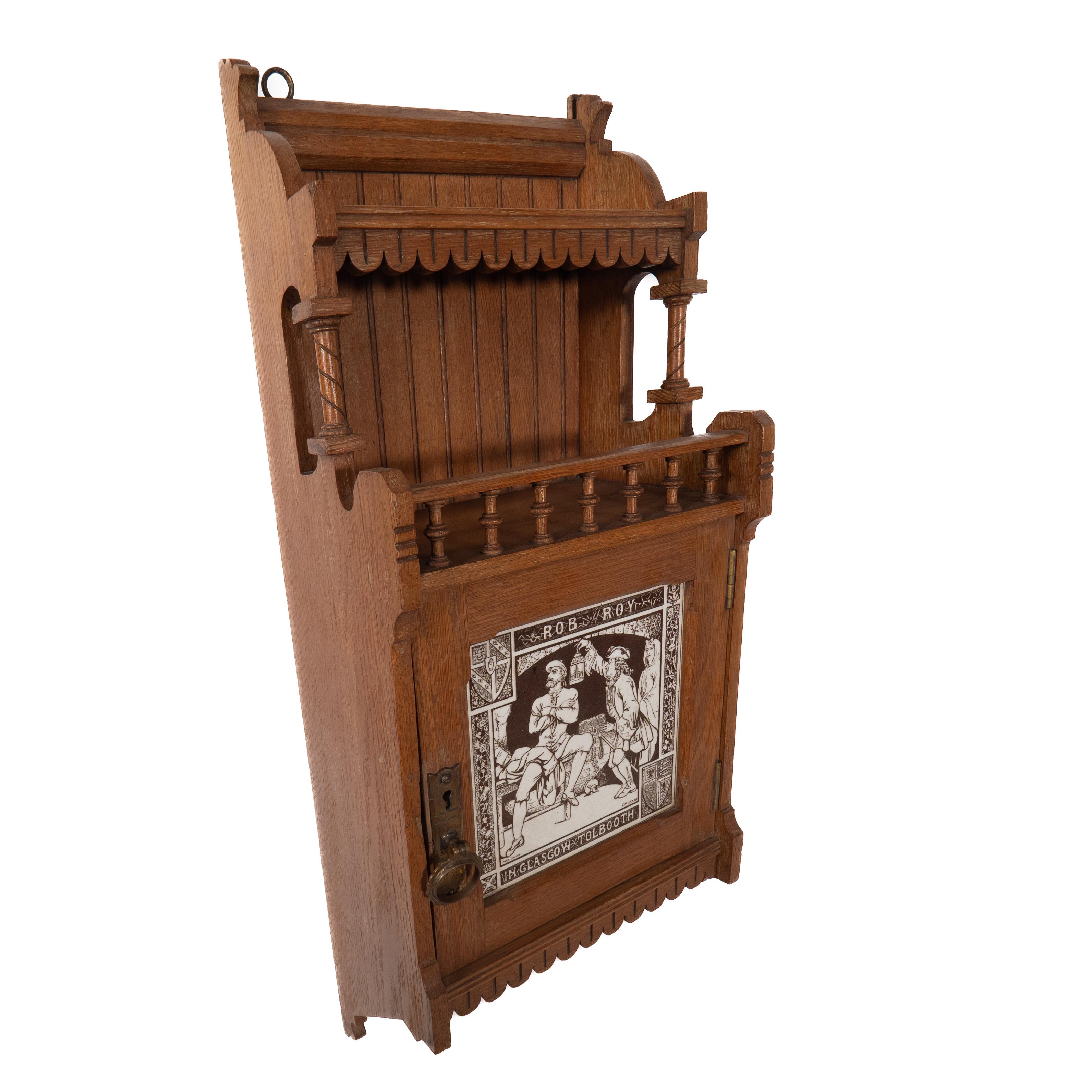 A reformed Gothic hanging bookcase, in the style of E W Godwin with a turned gallery and shelf, the door inset with a Minton 8 inch tile 'Rob Roy in Glasgow Tollbooth'.