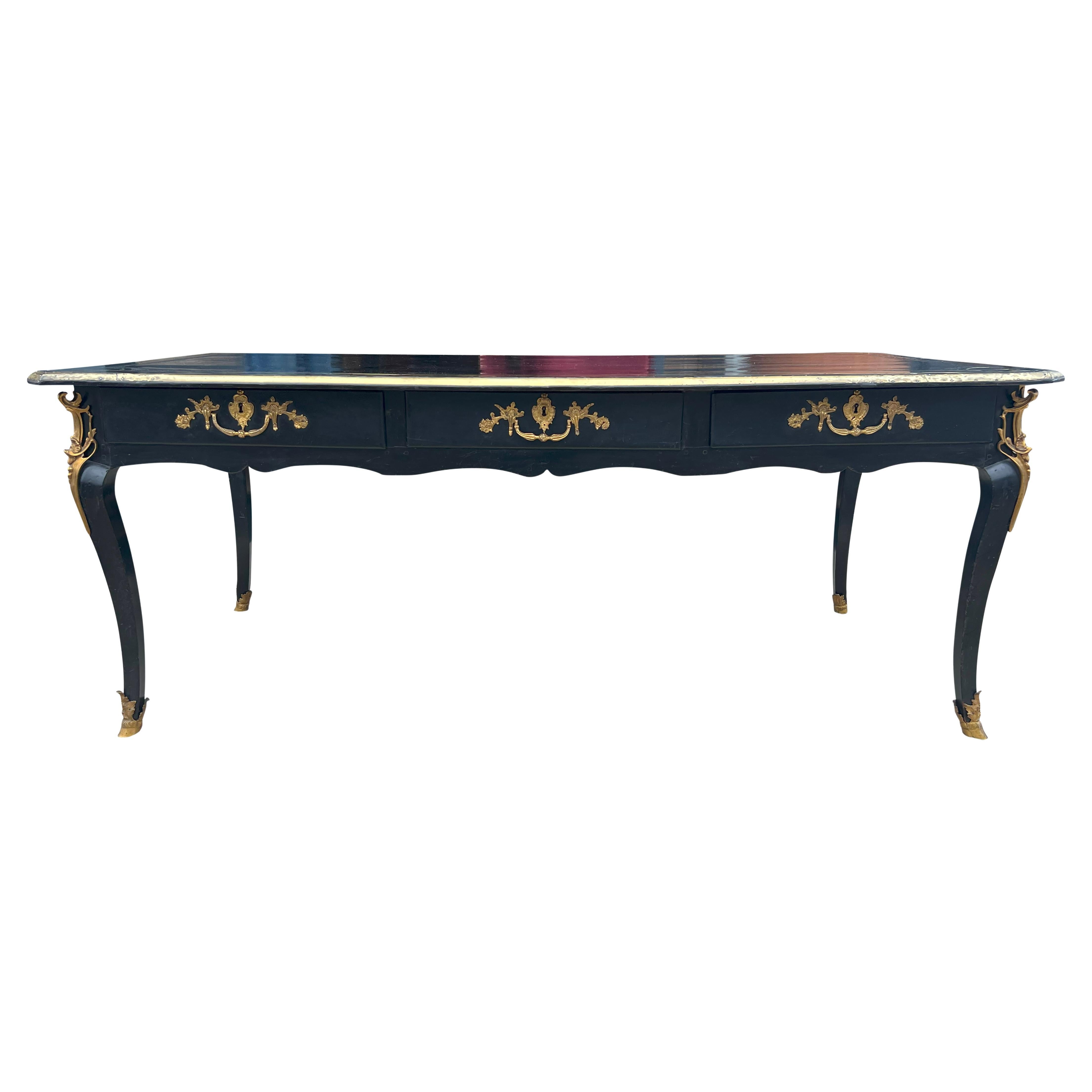With a rectangular top with inset black tooled leather surrounded by a brass inlaid ebonized border with brass edge. The frieze with three drawers with cast bronze handles and three false drawers on the reverse. Raised on cabriole legs and bronze