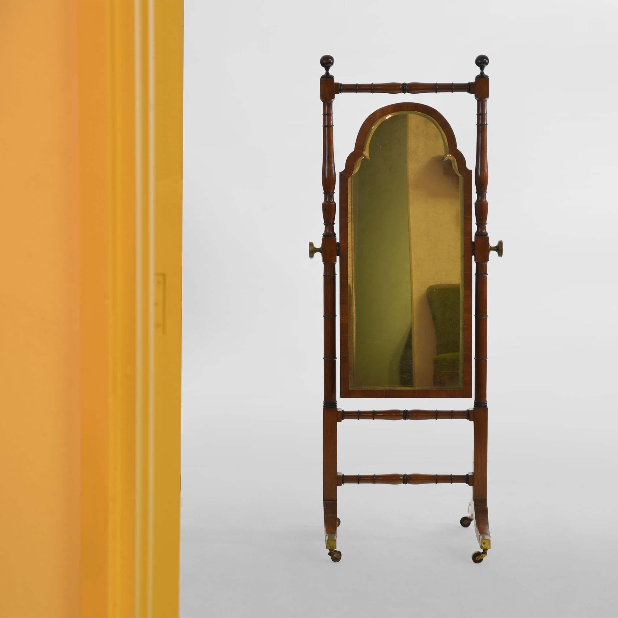 Regency mahogany cheval mirror, the turned frame with ball finials to trestle end supports with swept feet that terminate with brass box castors. The arched and bevelled mirror plate within a cross-banded frame with ebony stringing. The reverse is