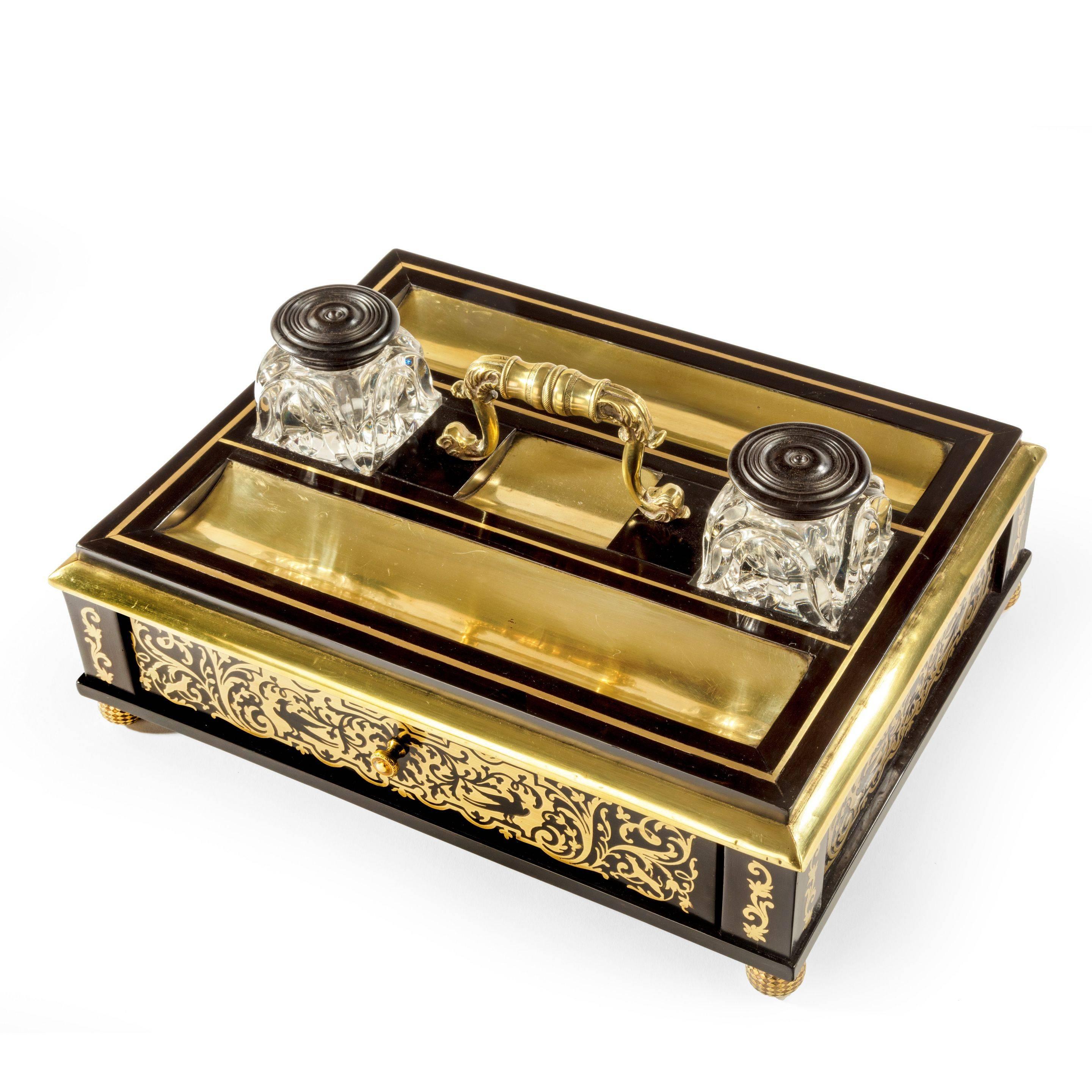 A Regency brass-inlaid ebony desk compendium of rectangular form with a
carrying handle flanked by cut glass inkwells between two pen trays, with a central drawer, set on bun feet, original key, an almost identical pen tray by W Leuchars was sold