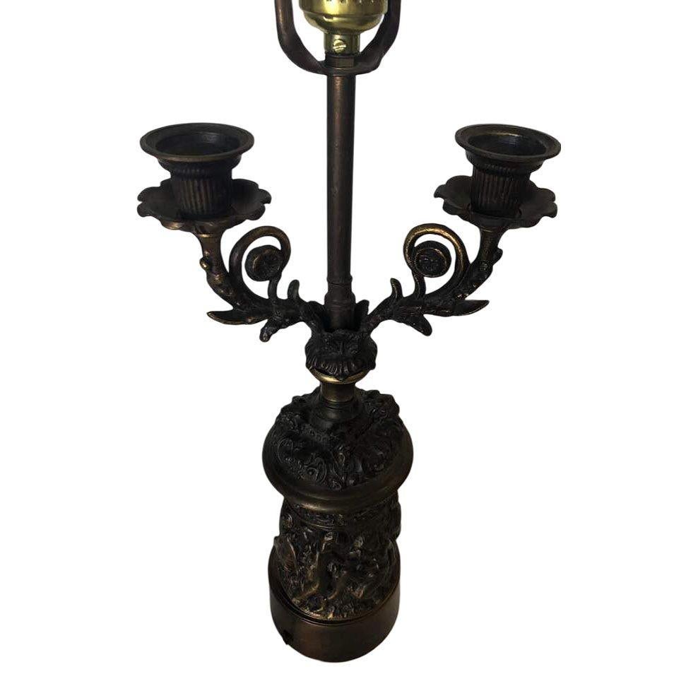 Neo-Classic Bronze Cherub Candelabra Table Lamp featuring a single light socket and Two Candleholders. The table lamp features a heavy relief of Cherubs wrapping around the base and scrolling patterns along the two decorative candle stick