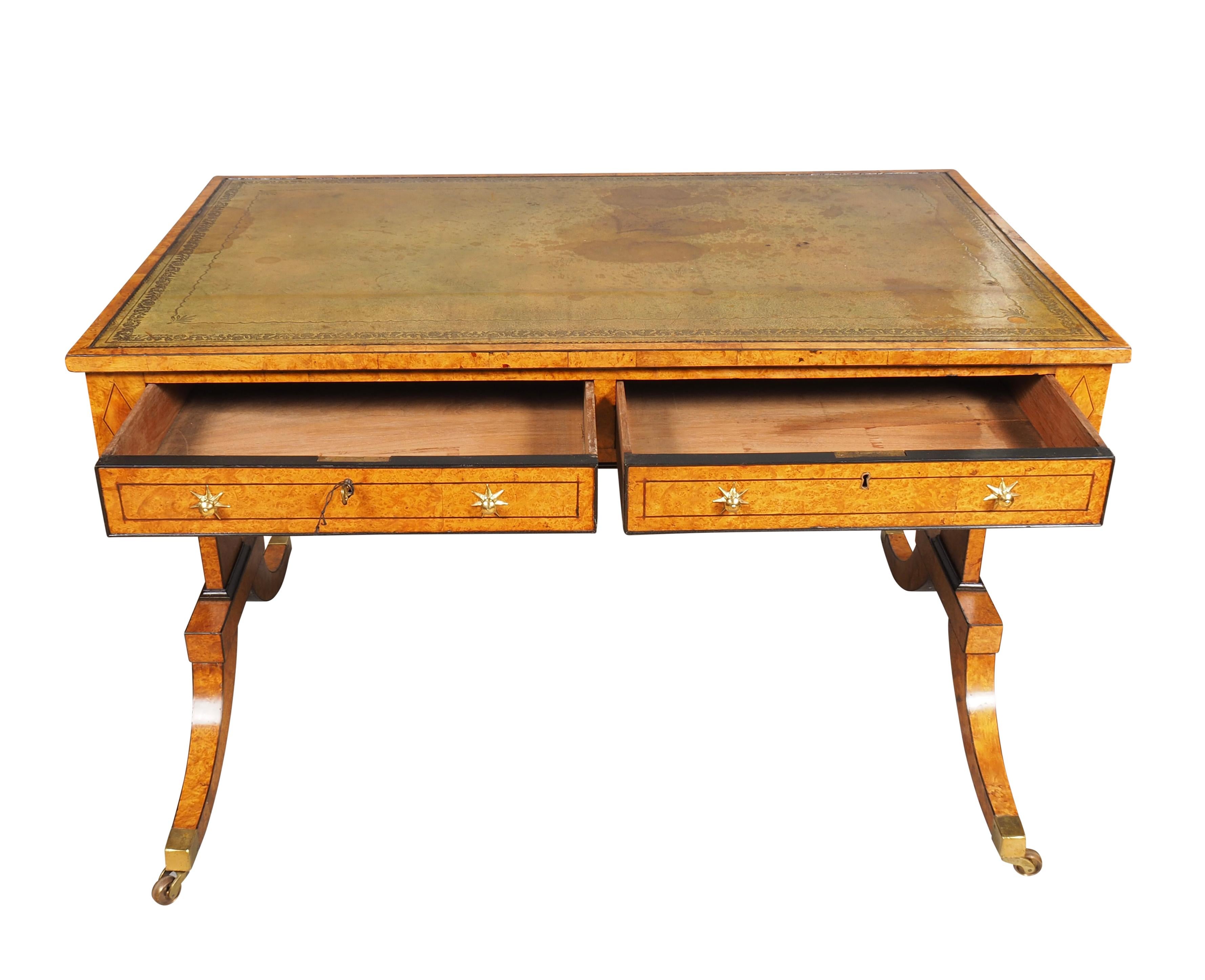 With a rectangular with an inset green leather top and banded edge. The frieze with two drawers and two opposing false drawers all with Gillow style star form brass knobs. Trestle support and saber legs with casters. Leather is badly stained and can
