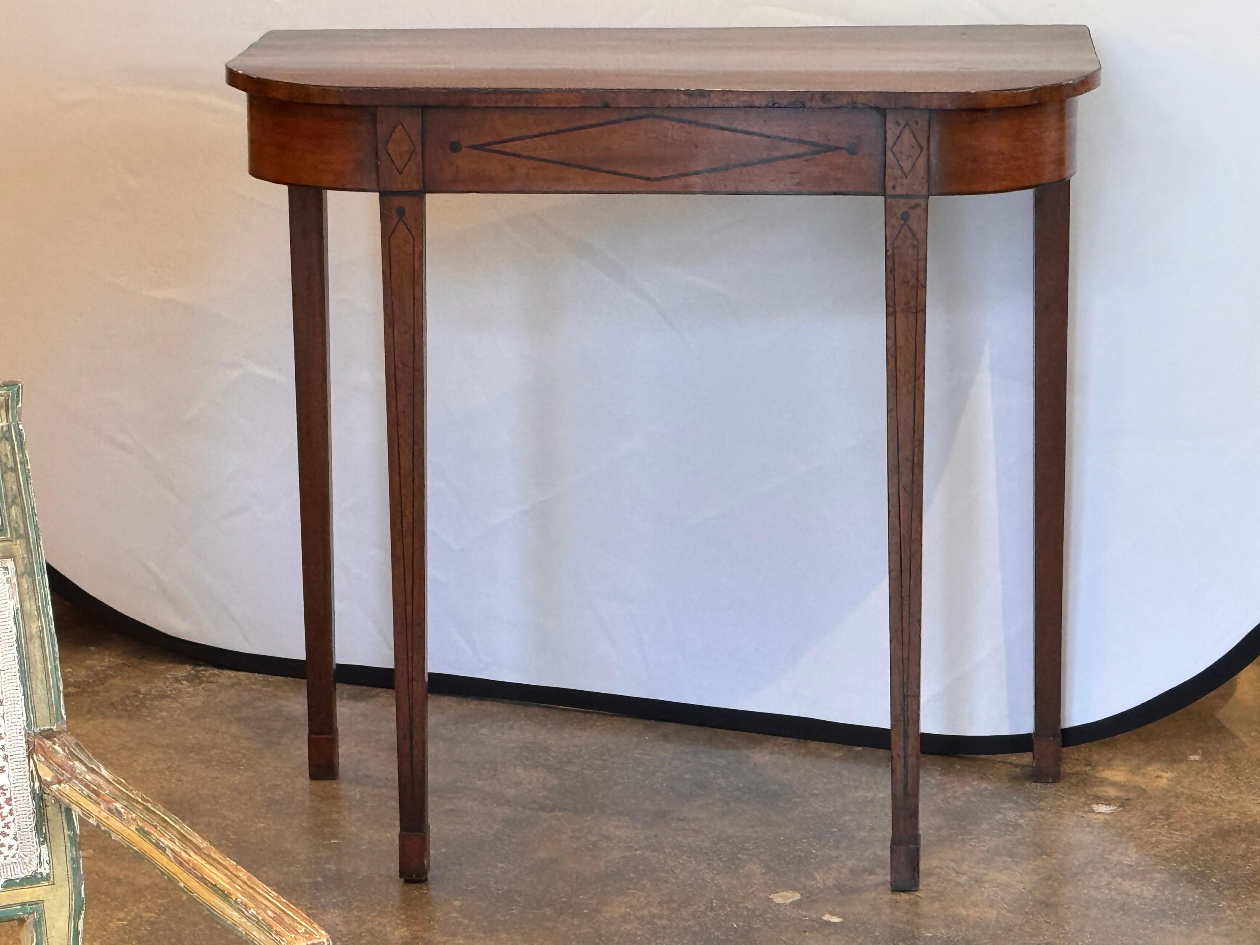 Consola estrecha con tablero en forma de arco y patas cónicas, adornada con delicadas incrustaciones decorativas a lo largo del faldón y las patas. Las incrustaciones lineales y de diamantes aportan un refinado toque neoclásico típico de la época de