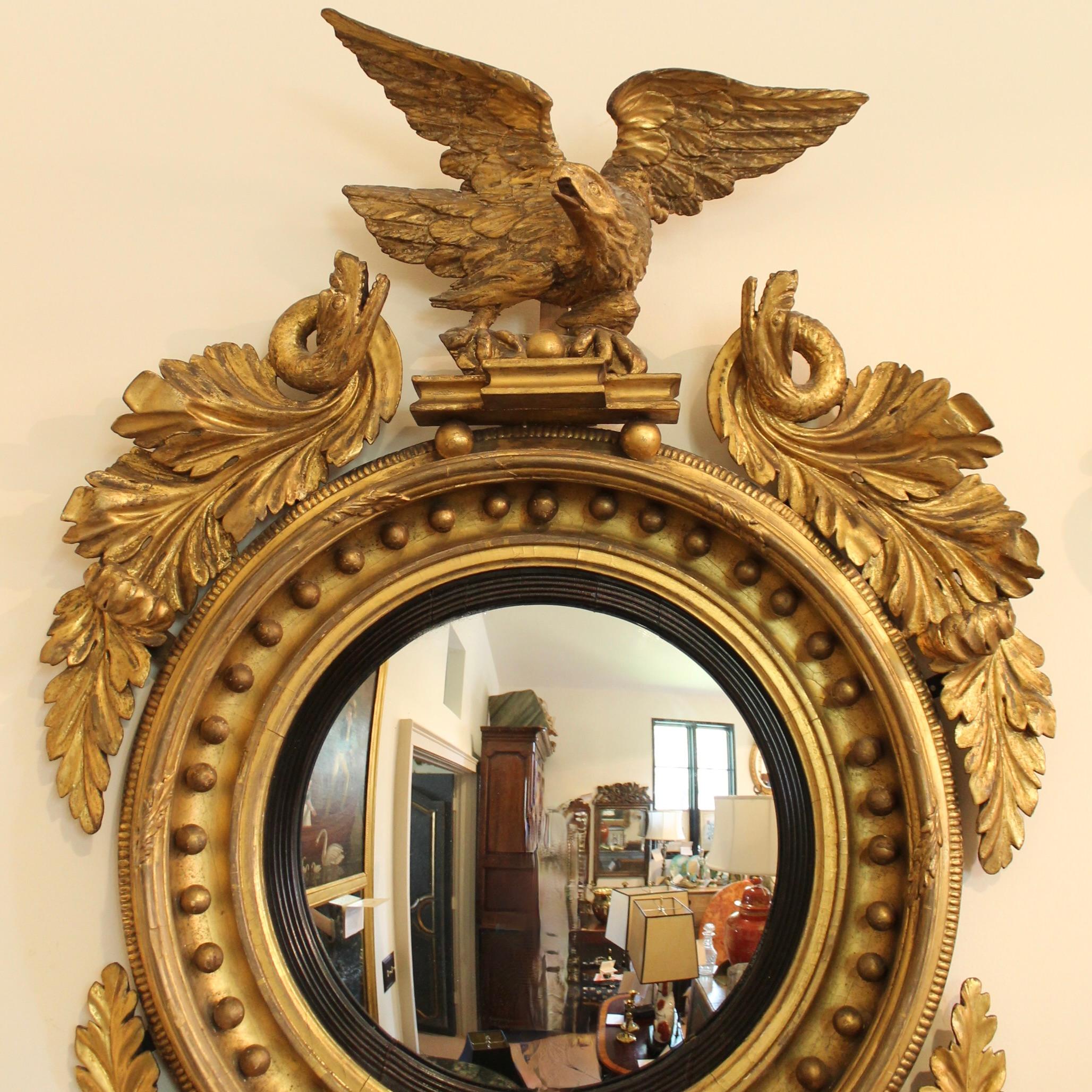 A sizable antique giltwood convex mirror carved in an assured hand with an eagle on top and swirls of carved and gilded acanthus foliage at the sides, the uppermost transforming into fantastical sea serpents. A tautly carved scallop shell