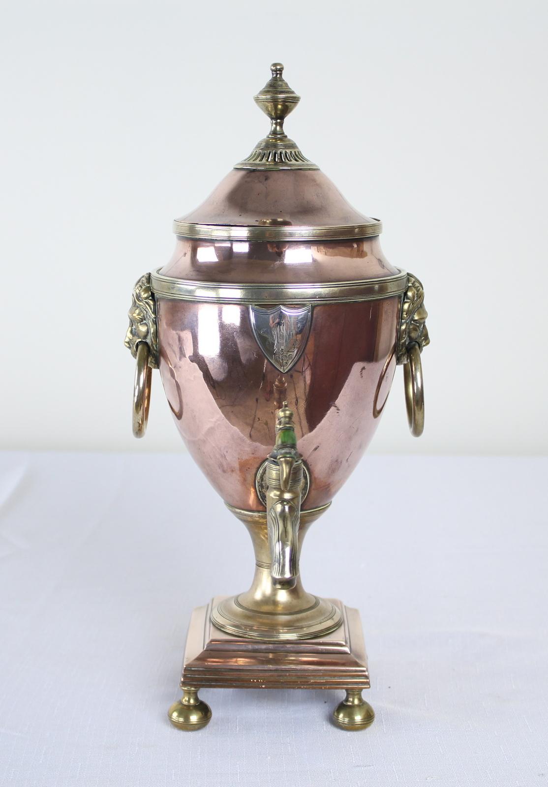 Un petit samovar ou une urne en cuivre sur un socle surélevé. Poignées en laiton en forme de tête de lion de chaque côté et couvercle à soulever avec poignée finale tournée. Petite patte gravée en argent au-dessus du bec.