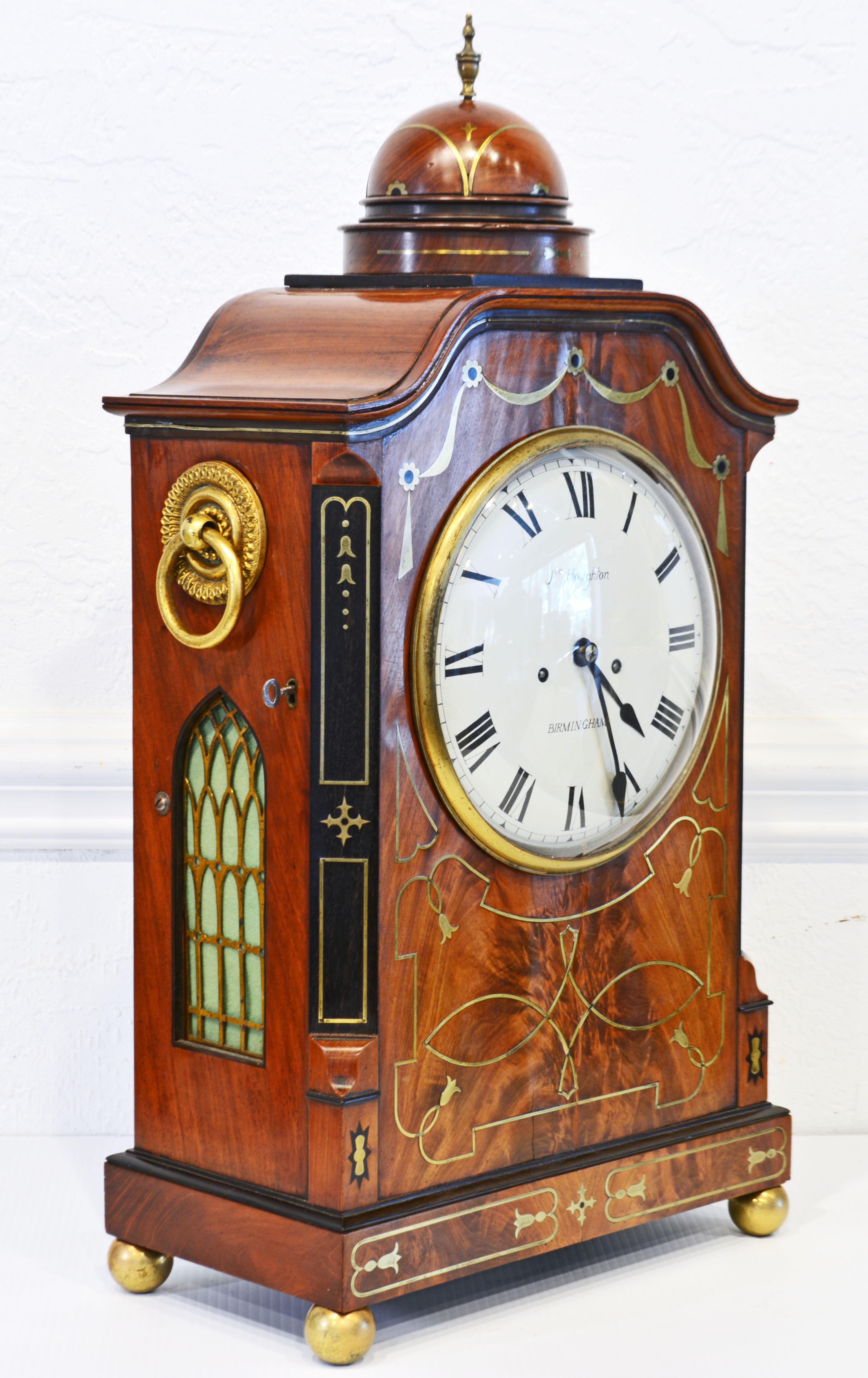 This elegant Regency mahogany Mantle clock features a domed top with brass finial on a bell shaped case top. The case sports a brass framed glass door with lock opening up to a white dial with Roman numerals and inscribed with maker's name and