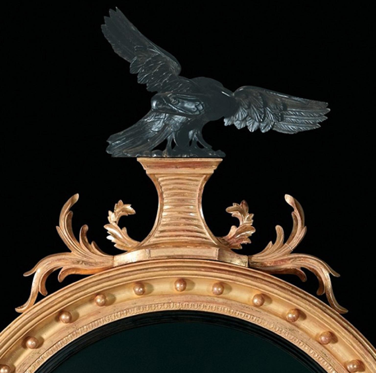 Un miroir convexe en bois doré sculpté de style Regency, avec un cadre moulé ébonisé et décoré de boules. Le cimier en forme d'aigle ébonisé repose sur un socle effilé et cannelé, flanqué de feuilles d'acanthe enroulées et d'un tablier sculpté de