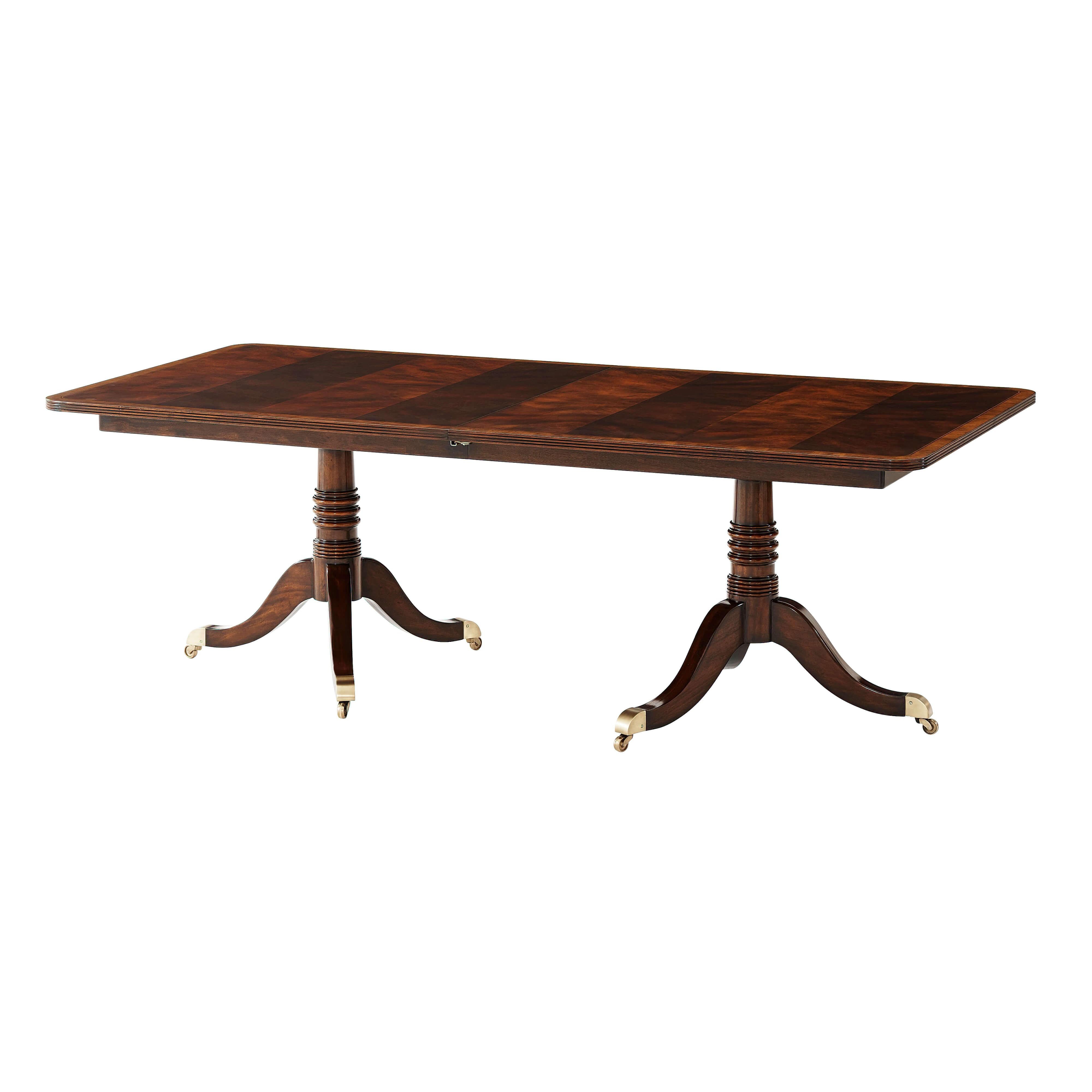 An English Regency style figured mahogany veneered dining table, the rectangular reeded edge and satinwood banded top extending to accommodate one additional leaf, on two gun-barrel turned column pedestals with down swept legs terminating in brass