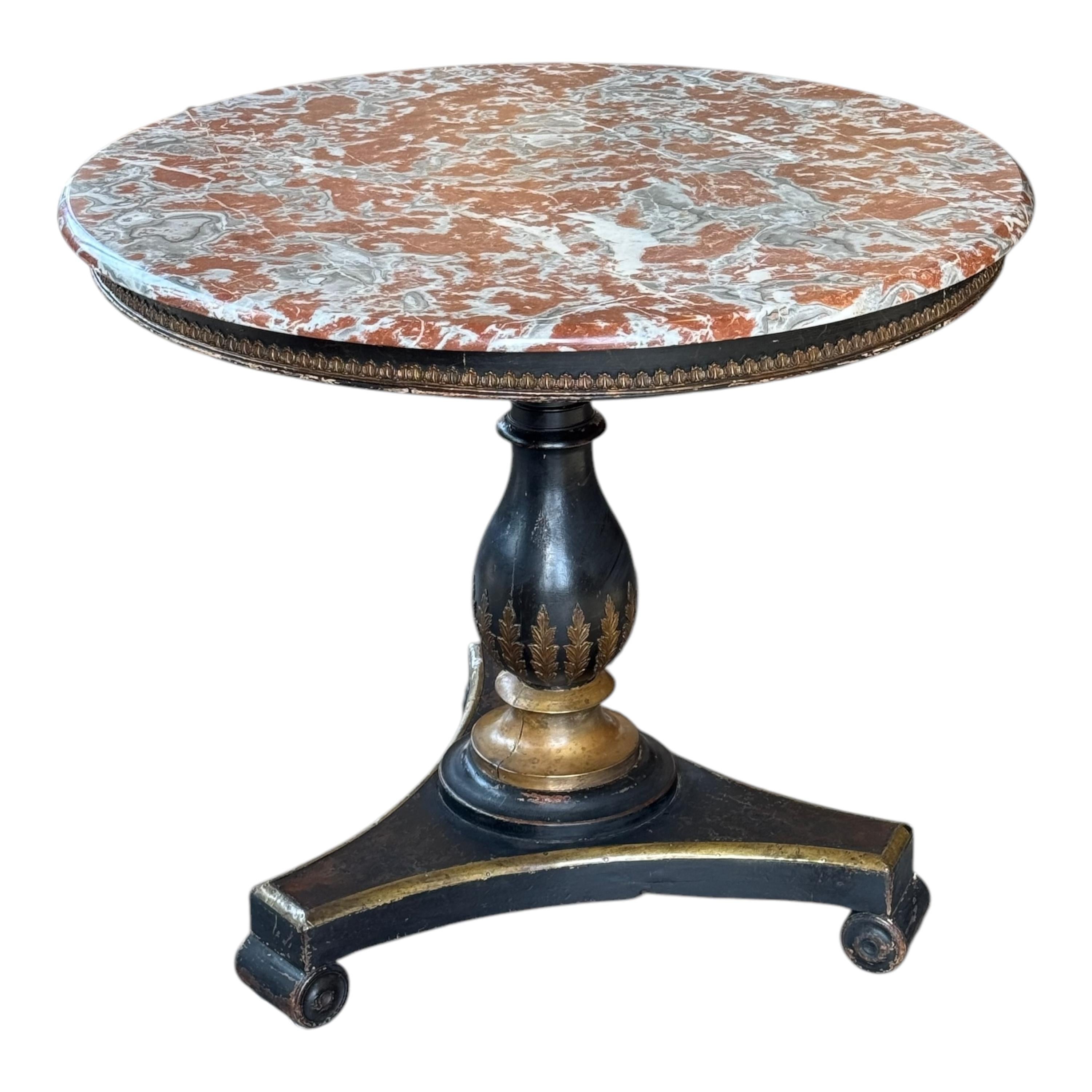 Regency-style center table featuring a striking round marble top with rich veining. The ebonized base is adorned with gilt detailing, including finely painted foliate motifs on the urn-shaped pedestal. The tripartite base is trimmed with accents. A