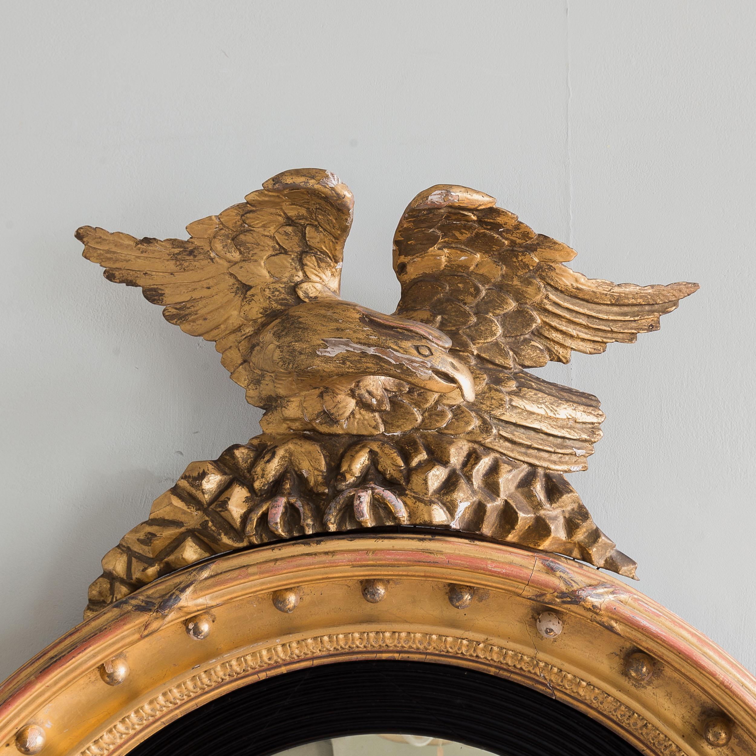 A large Regency gilded convex mirror, early nineteenth century, surmounted by cresting of an eagle perched and poised on rocks, the circular frame with balls and delicate lambrequin mould, a generous reeded and ebonised slip within and surrounding