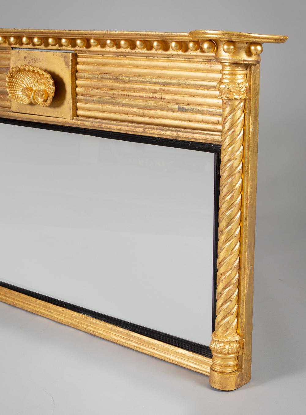 A Regency carved giltwood overmantle mirror, the molded cornice with protruding round corners, above ball spacers and a ribbed frieze decorated with a carved three-dimensional shell, the central mirror plate with ebonized reeded slip molding is